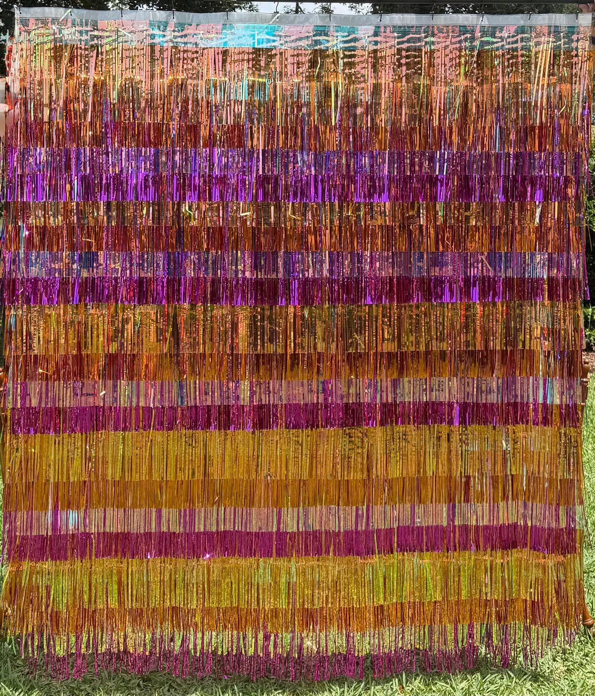 A vertical tinsel fringe curtain or door hanging made of metallic strands in horizontal bands of purple, orange, gold, and green, photographed outdoors on grass with trees visible in the background.