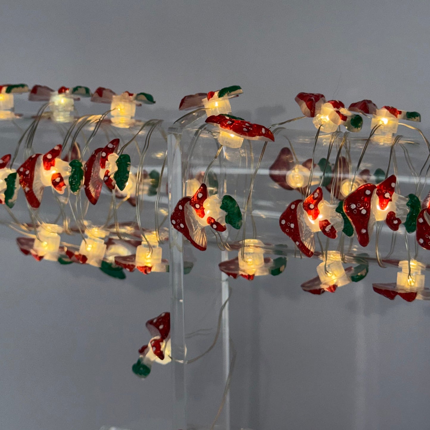 Christmas tree string lights wrapped around a clear spiral tree stand, featuring red ornaments with white trim and green accents, glowing with warm white lights against a plain gray background.