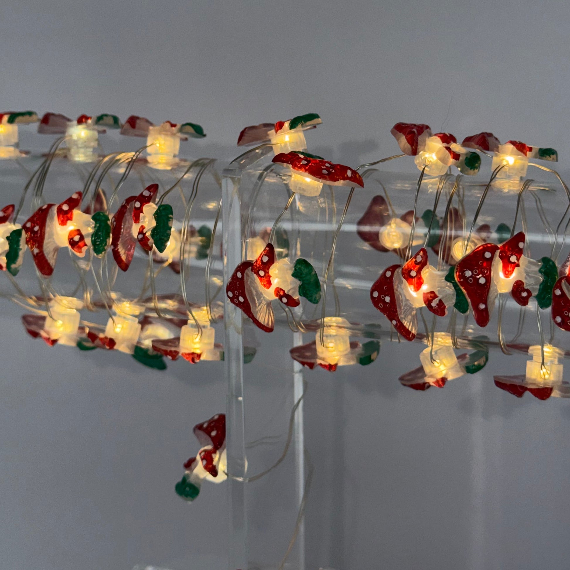 Christmas tree string lights wrapped around a clear spiral tree stand, featuring red ornaments with white trim and green accents, glowing with warm white lights against a plain gray background.
