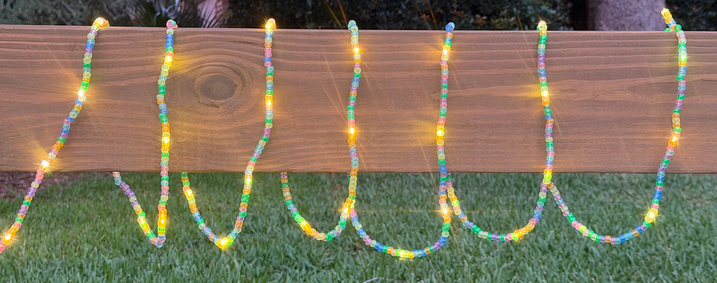 Multicolor beaded string lights draped in wavy vertical lines over a wooden fence on a grassy lawn at dusk.
