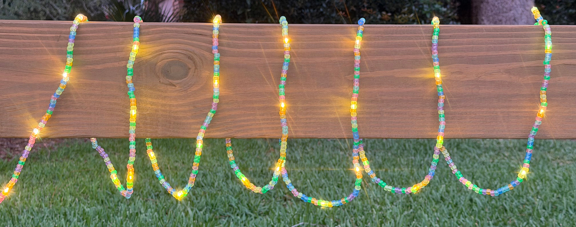 Multicolor beaded string lights draped in wavy vertical lines over a wooden fence on a grassy lawn at dusk.