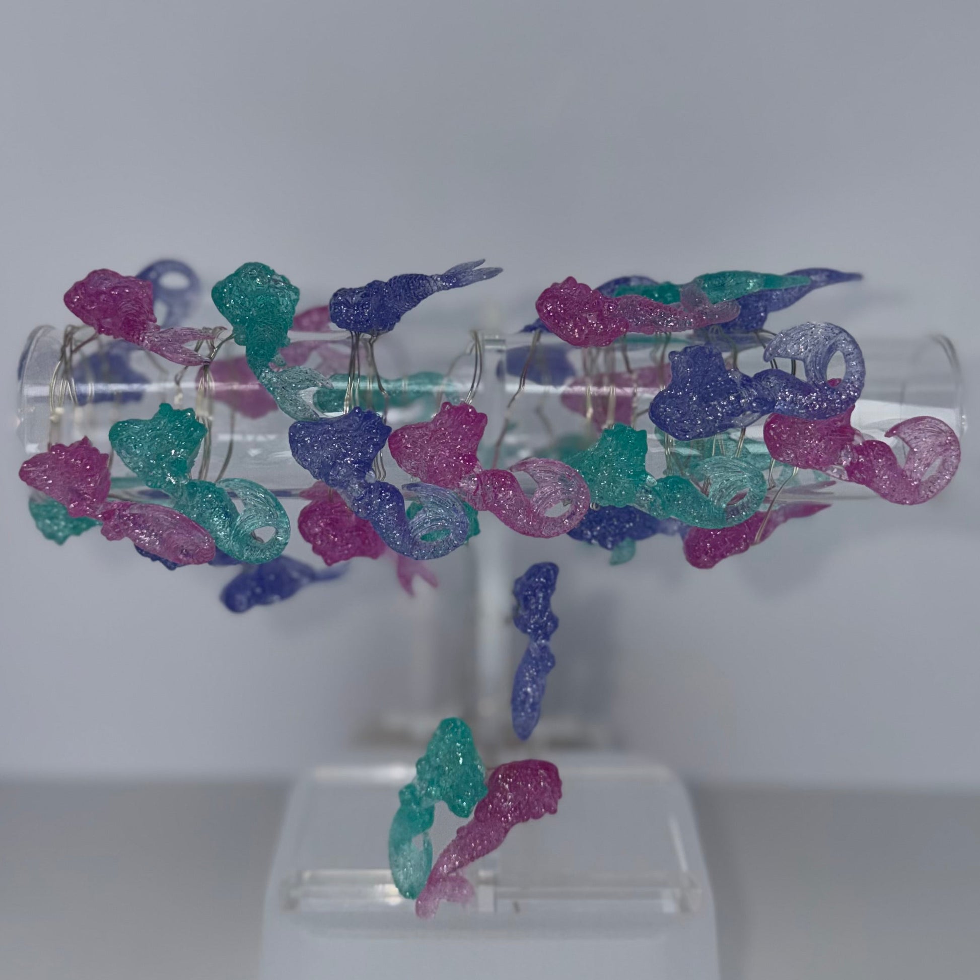 A clear acrylic jewelry display stand holding multiple strands of small, glittery seed bead necklaces in purple, teal, and pink, set against a plain light gray background.