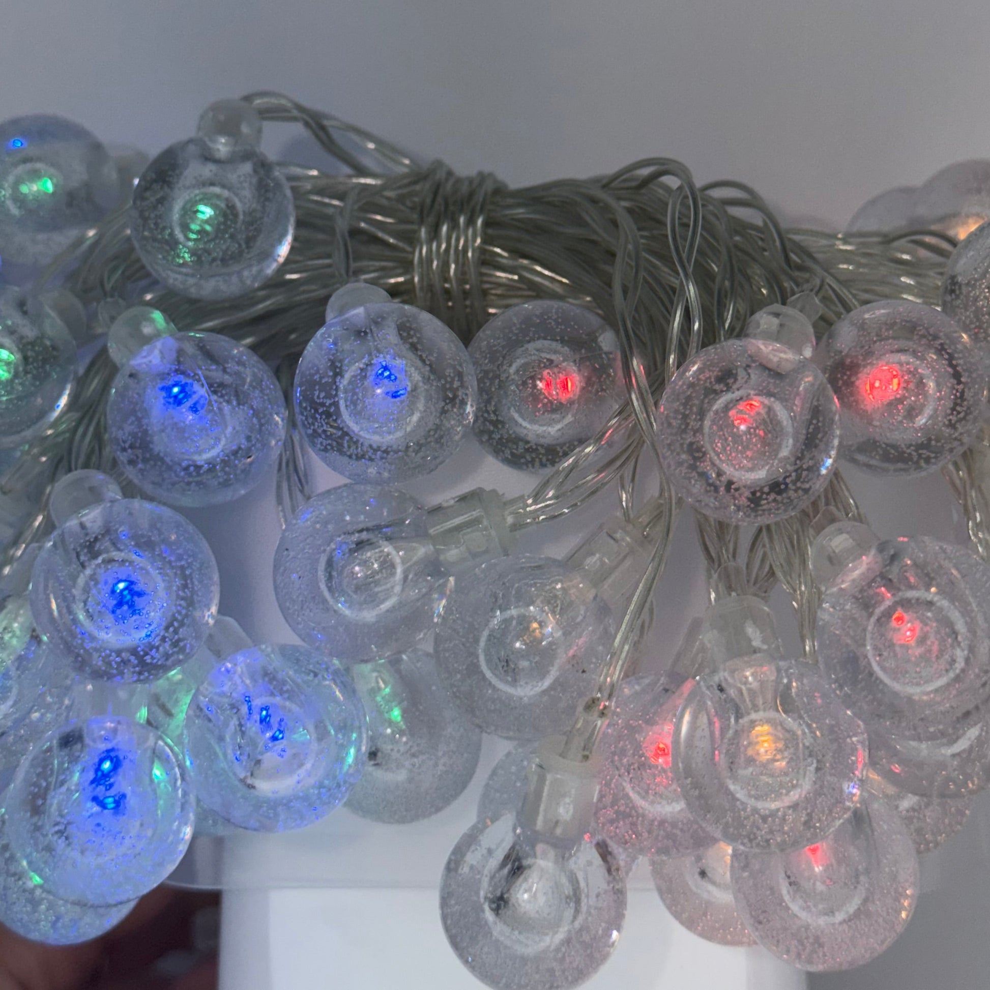 A close-up of a bundle of clear plastic bubble string lights glowing with multiple colors, including blue, green, red, and orange, wrapped in a silver wire. A hand is holding the right side of the light bundle against a plain light gray background.