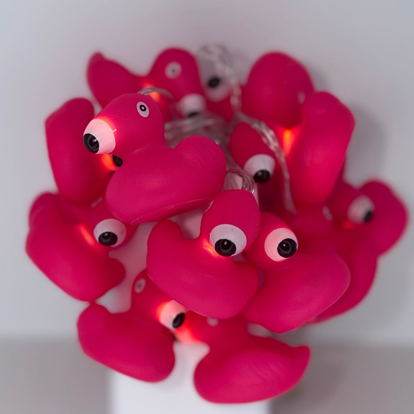 A cluster of pink flamingo-shaped string lights with glowing warm white bulbs, displayed on a white stand against a plain light gray background.