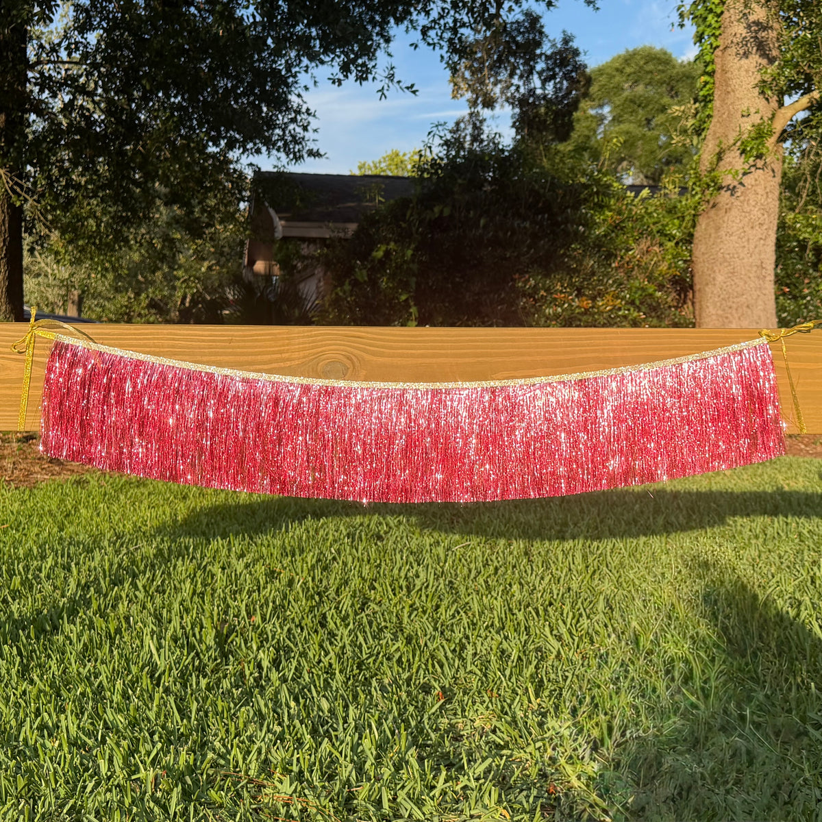 A pink metallic tinsel fringe banner hanging outdoors across a wooden fence on a green grass lawn, with trees and a house in the background.