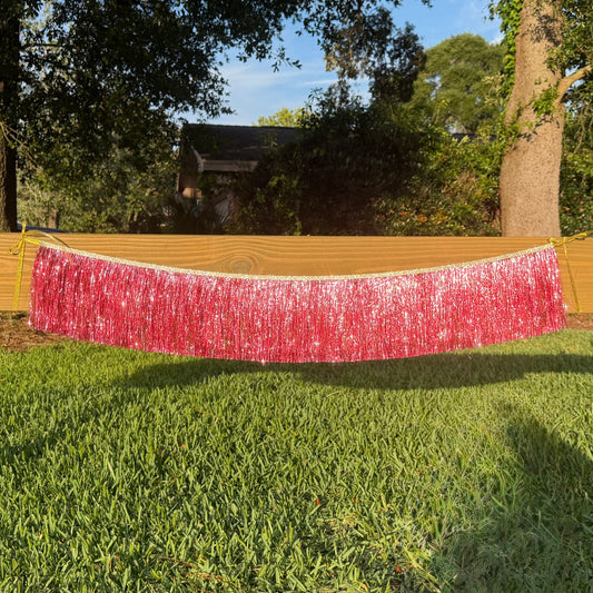 A pink metallic tinsel fringe banner hanging outdoors across a wooden fence on a green grass lawn, with trees and a house in the background.