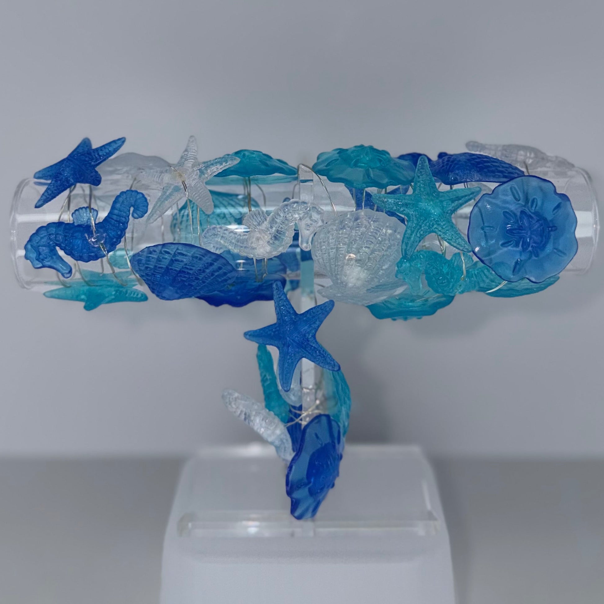 A blue and clear glass sculpture of sea life, including starfish, seahorses, and jellyfish, mounted on a white plastic display stand against a neutral gray background.