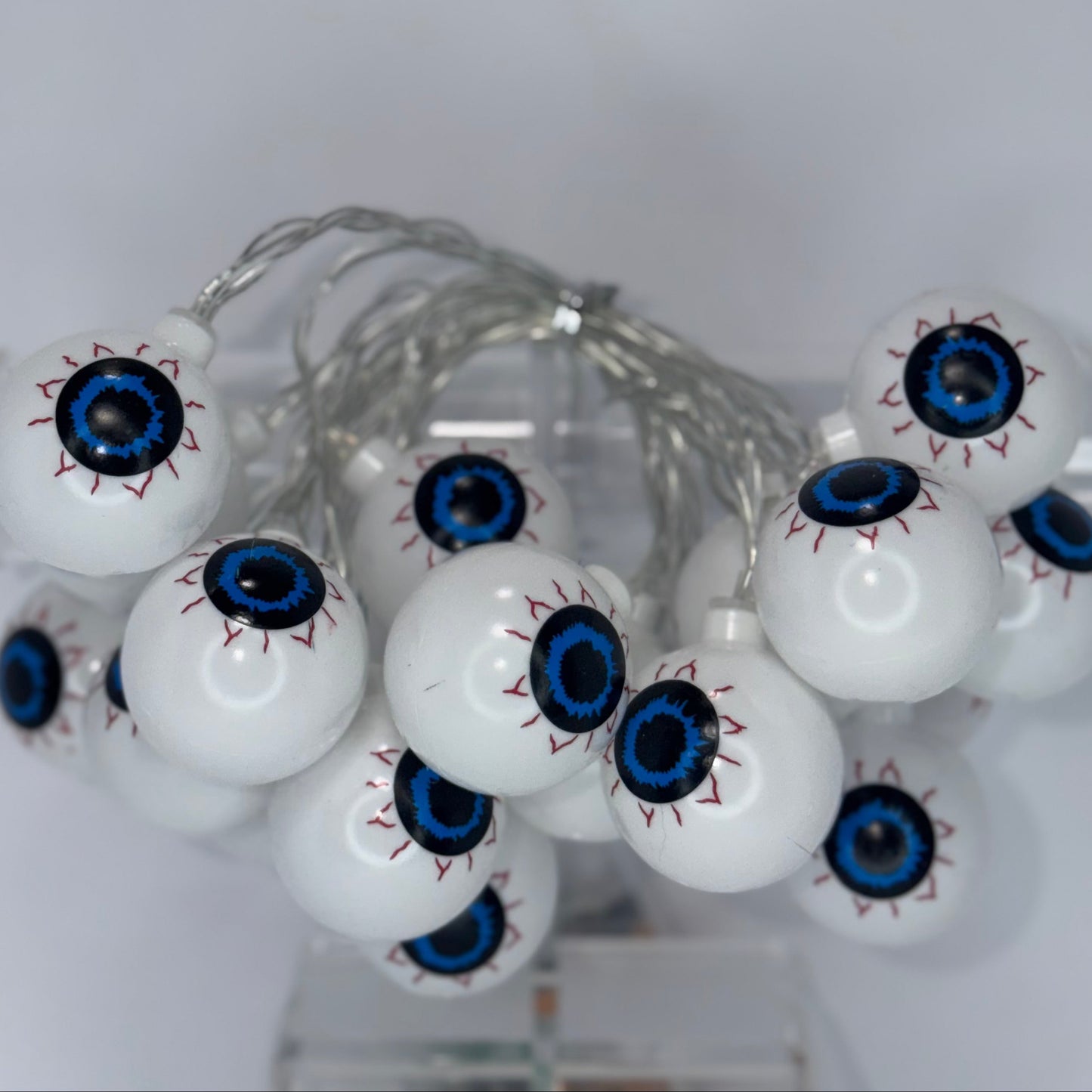 Glowing eyeball string lights with white plastic spheres, detailed blue irises, and red stitched details on a clear wire, displayed on a clear stand against a light gray background.