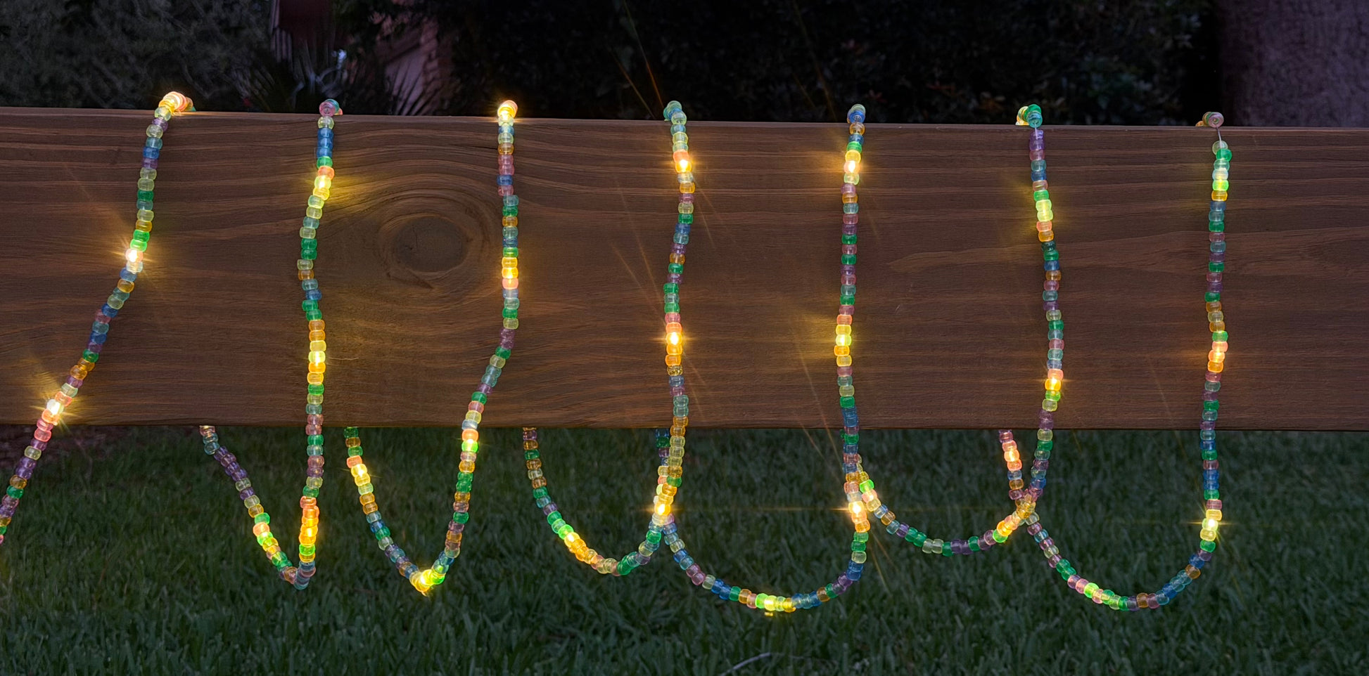 Multicolor LED bead garland lights draped over a wooden fence on a lawn at night.
