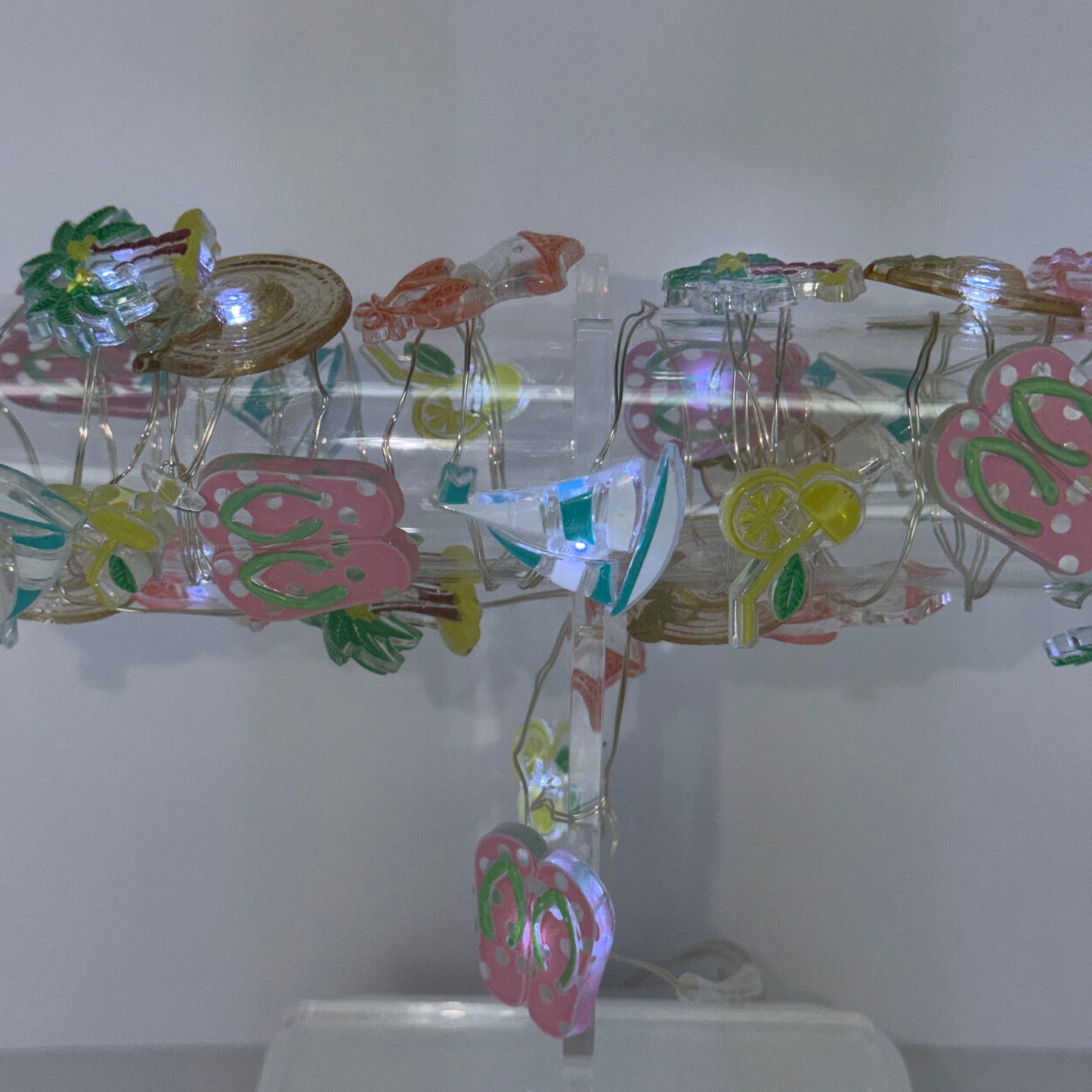 A clear acrylic hanging display stand decorated with colorful paper cutouts, small green leaves, and a butterfly, shown against a plain light gray wall.