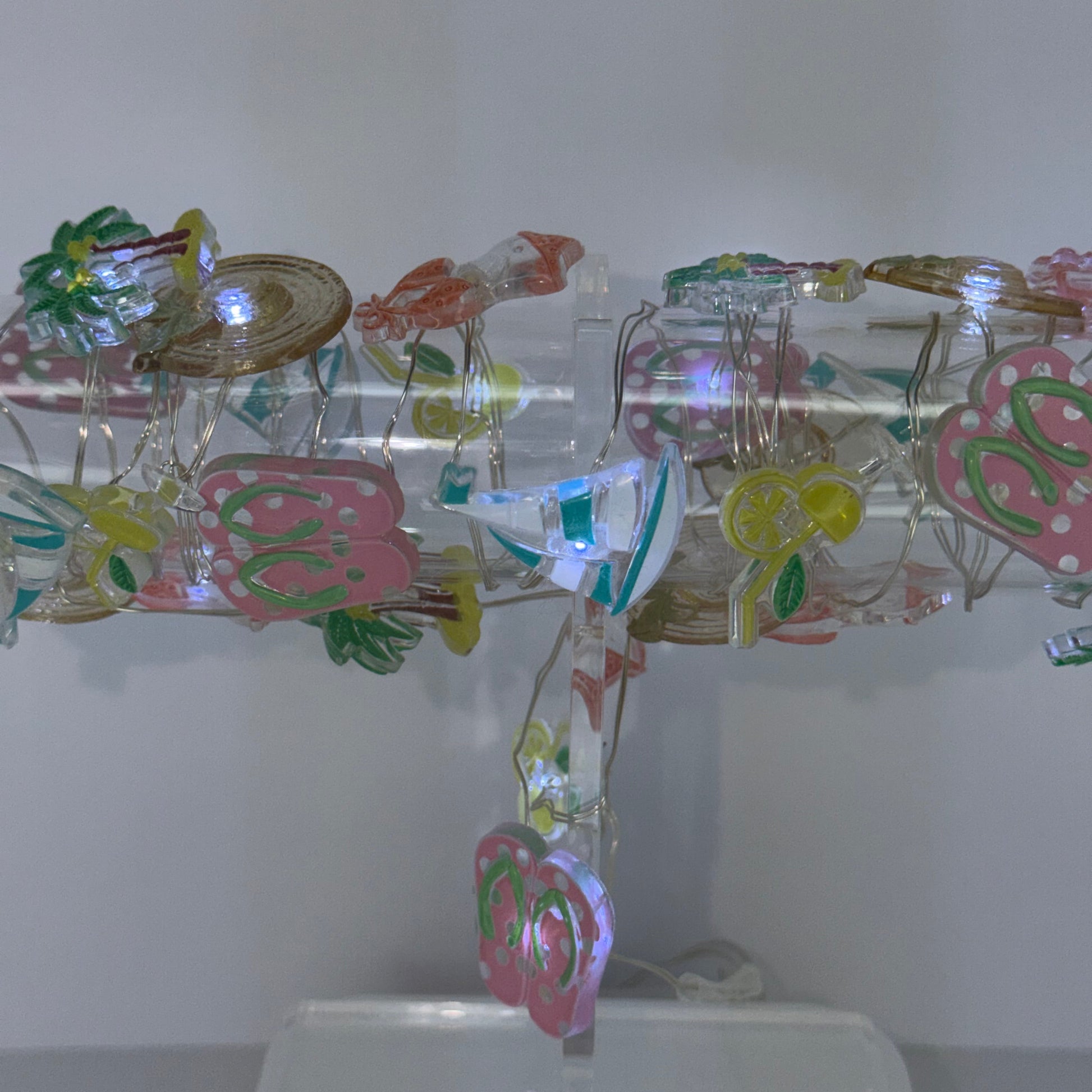 A clear acrylic hanging display stand decorated with colorful paper cutouts, small green leaves, and a butterfly, shown against a plain light gray wall.