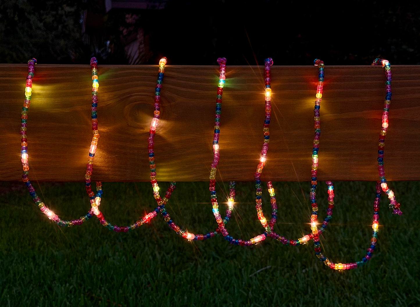 Multicolor beaded string lights draped over a wooden fence on a dark grassy lawn at night.
