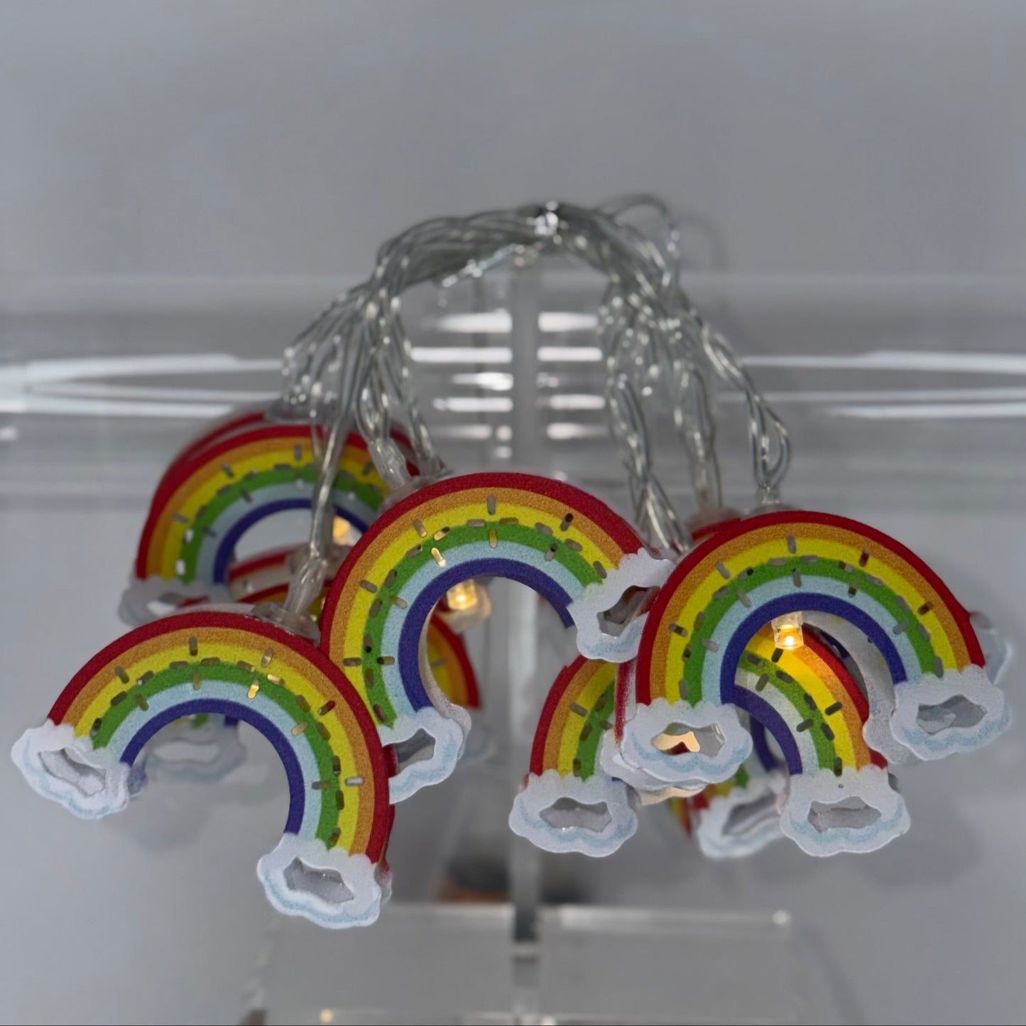 Rainbow LED string lights shaped like rainbows with white cloud accents, hanging from a clear acrylic display stand.