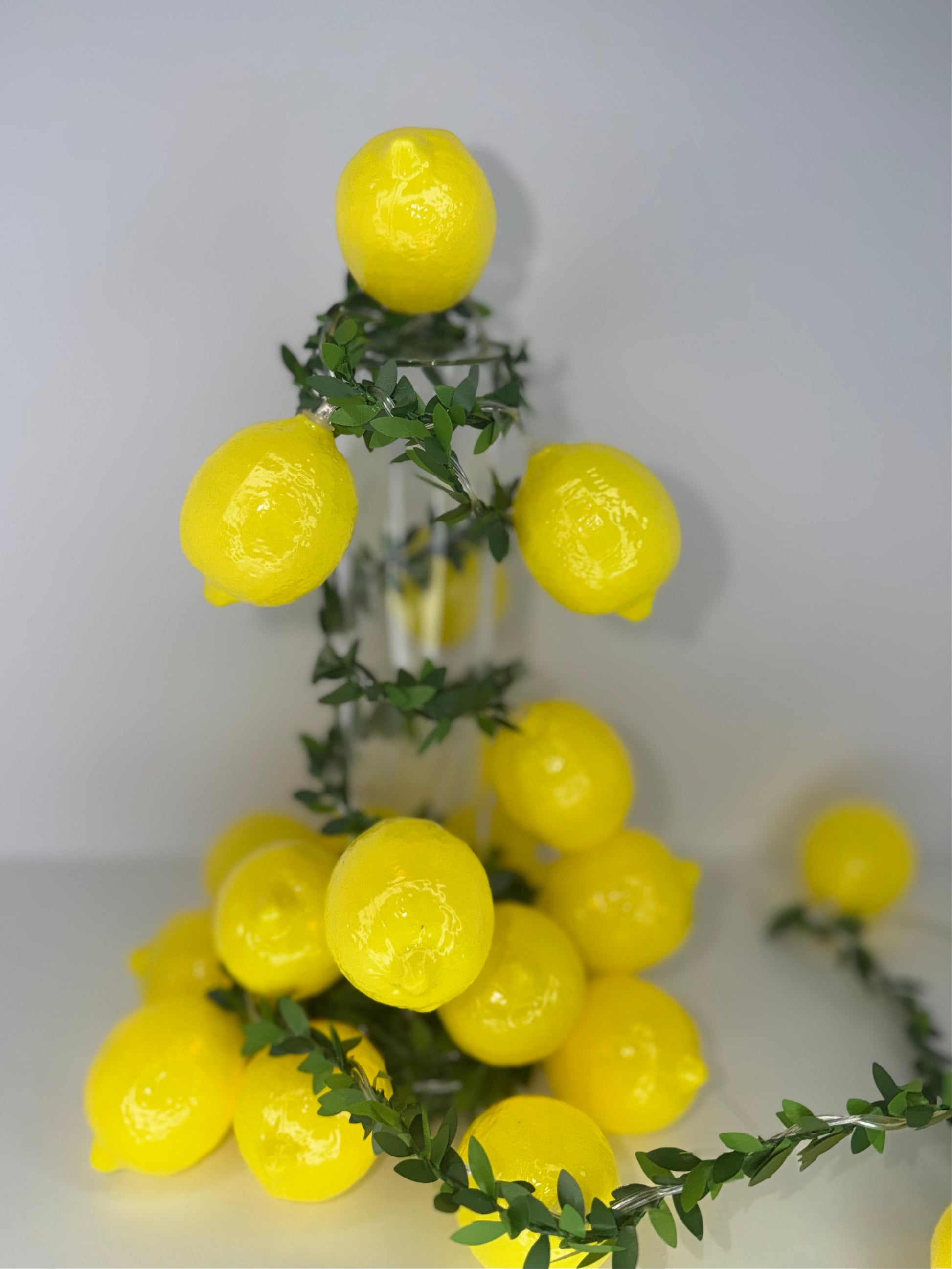 Yellow artificial lemon garland with green leaves draped over a clear glass vase, photographed against a plain light gray background.
