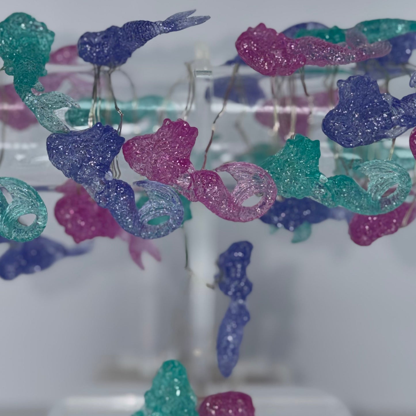 A close-up of a clear acrylic display stand holding multiple glittery butterfly-shaped earrings in translucent purple, teal, and pink tones against a soft light gray background.