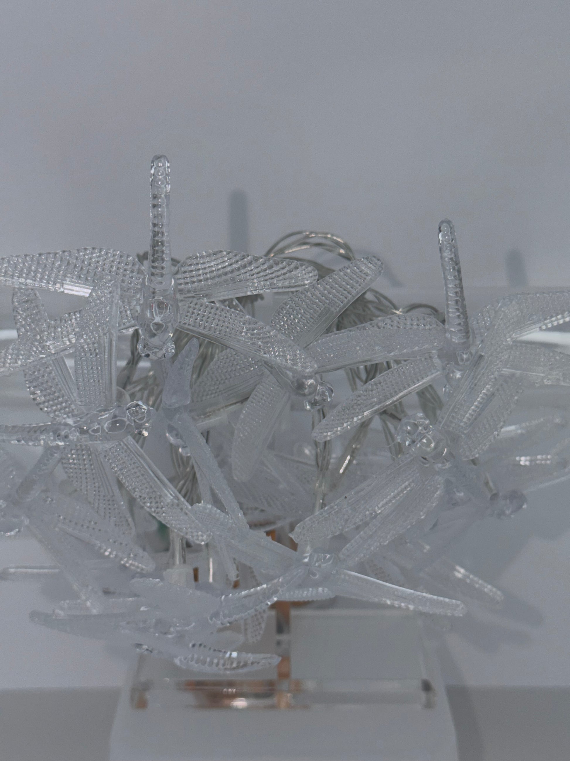 A cluster of clear, textured plastic dragonflies attached to a transparent stand against a plain light gray background.