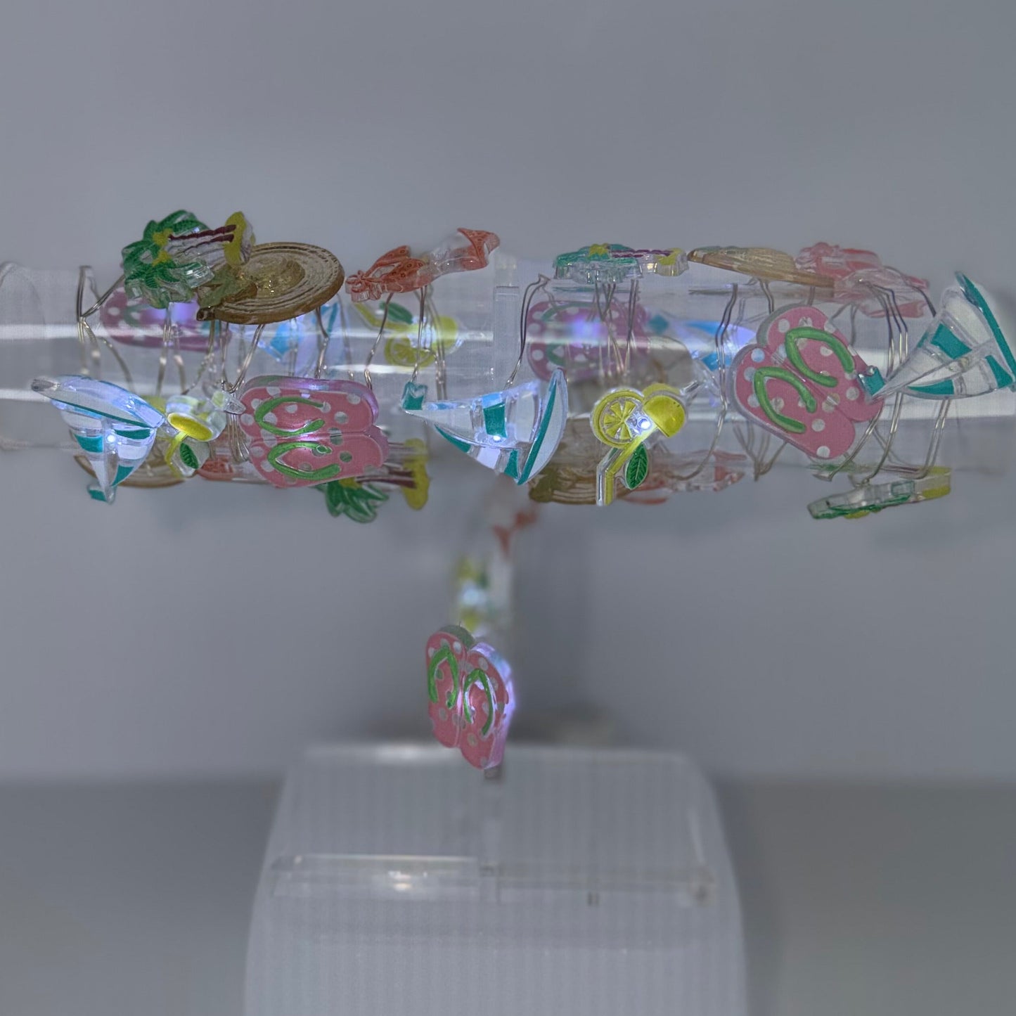 A clear cylindrical display tube mounted on a white plastic stand, filled with colorful iridescent foil sheets and small decorative elements, set against a plain light gray background.
