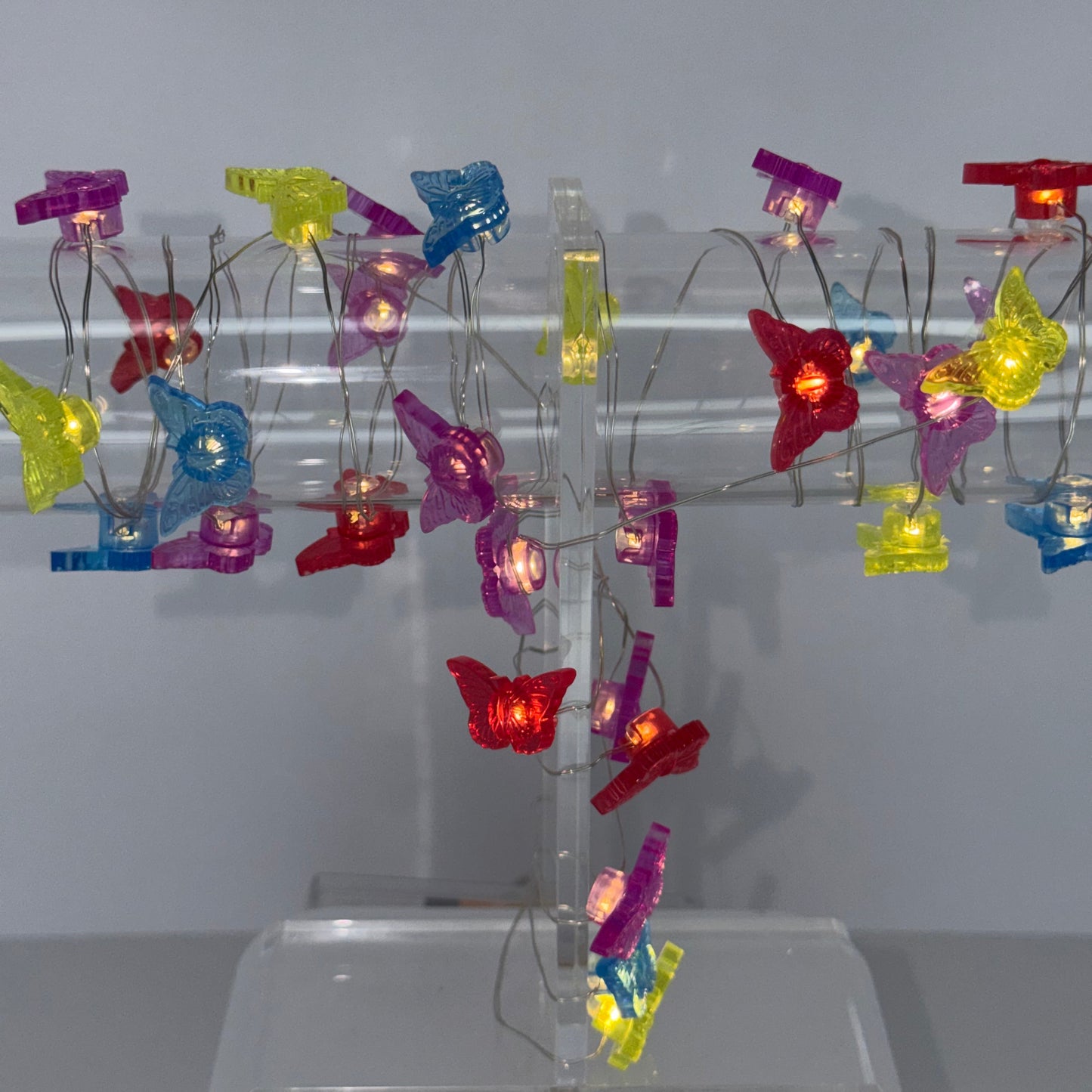 A decorative hanging light fixture made of a clear acrylic frame with many small translucent clip-style bulbs in red, yellow, blue, purple, and green, each glowing with warm white lights, shown against a plain light gray wall.