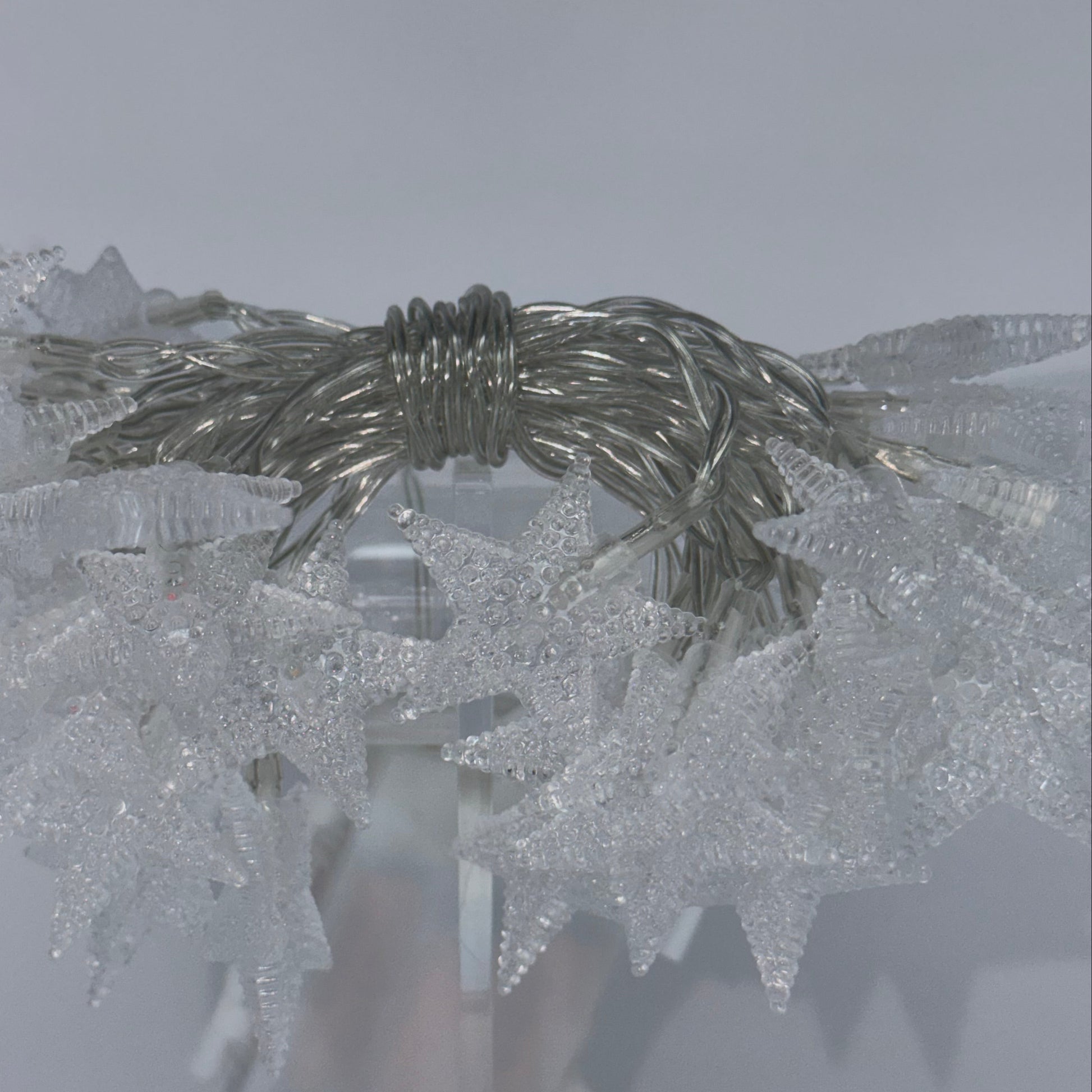 A close-up of a decorative silver wire wreath or garland wrapped with textured clear plastic mesh, shown against a pale gray background.