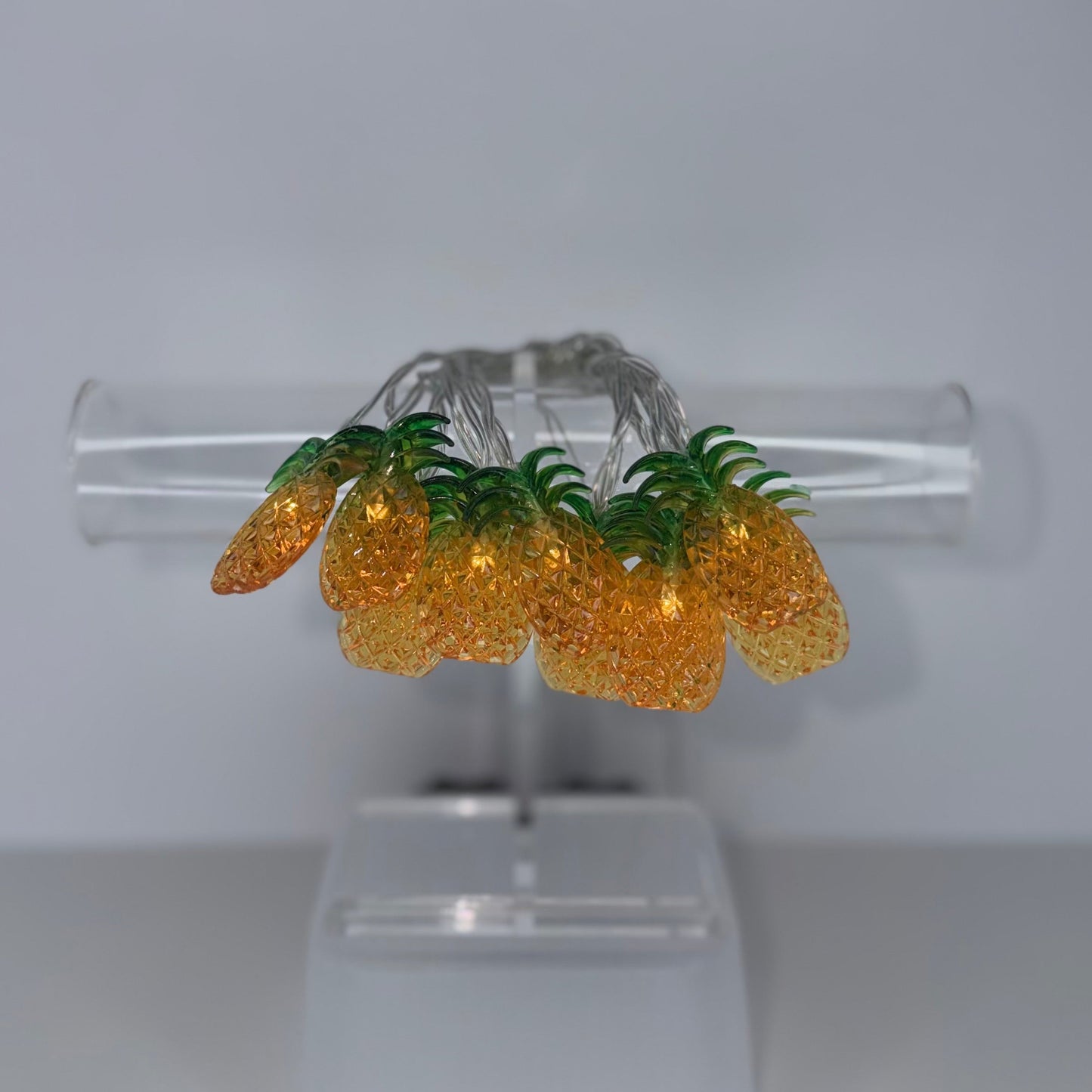 A pair of gold and green pineapple-shaped earrings displayed on a clear acrylic stand against a neutral gray background.