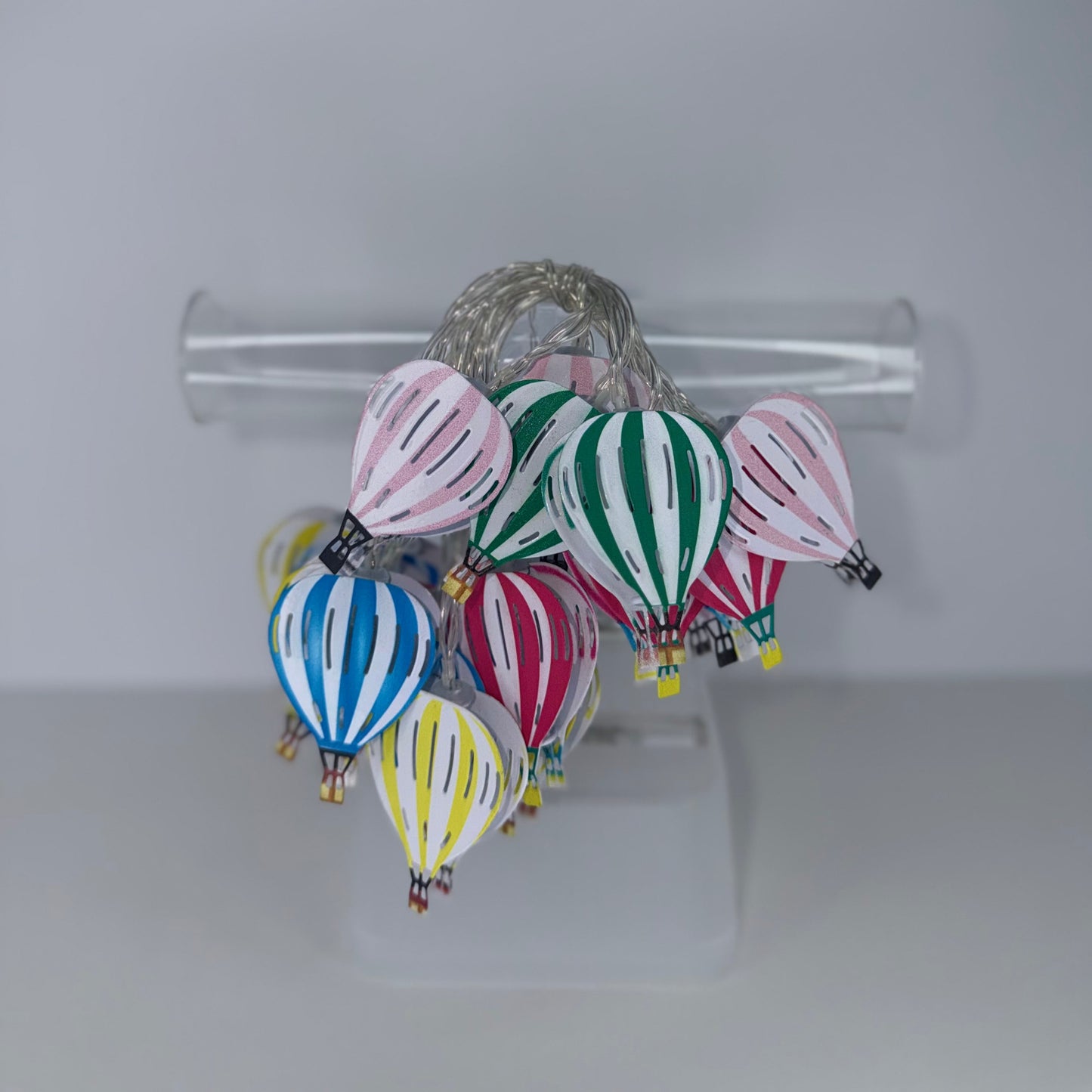 A cluster of small hot air balloon ornaments in multiple colors, including pink, green, blue, red, yellow, and white, hanging from a clear plastic stand against a plain light gray background.