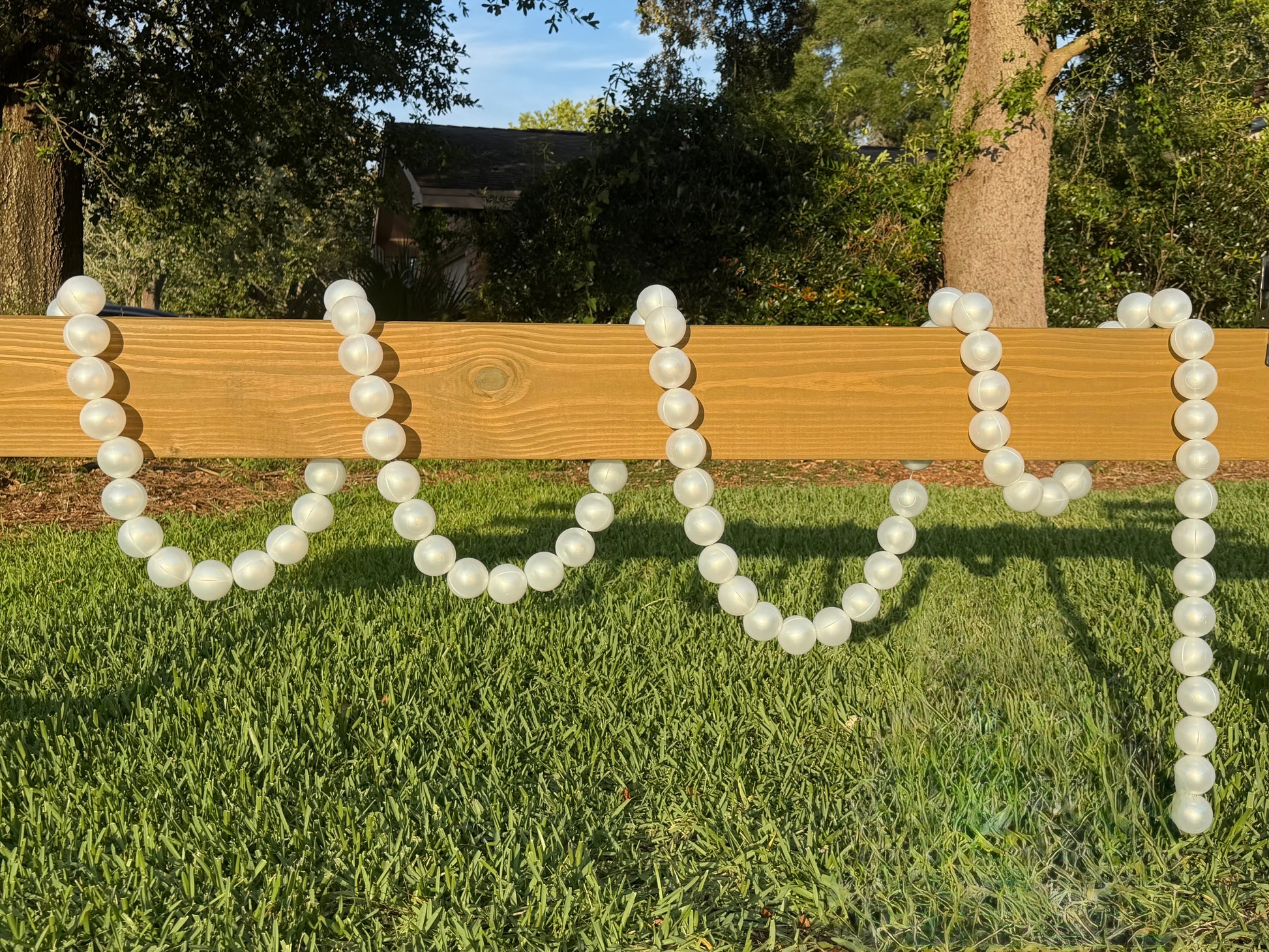 White faux-pearl beaded garland draped across a wooden fence on a green grass lawn with trees in the background.