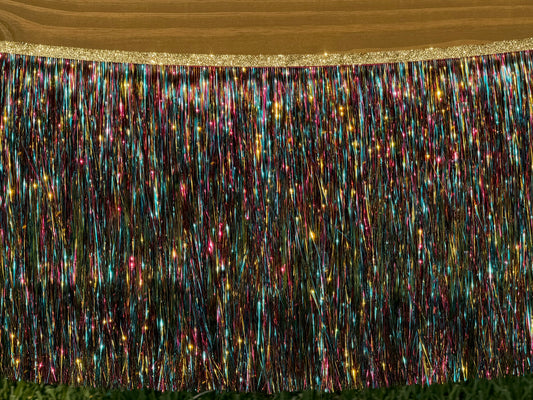 A close-up of a multicolor metallic tinsel fringe banner with a glittery gold top edge, shown hanging against a wooden background.