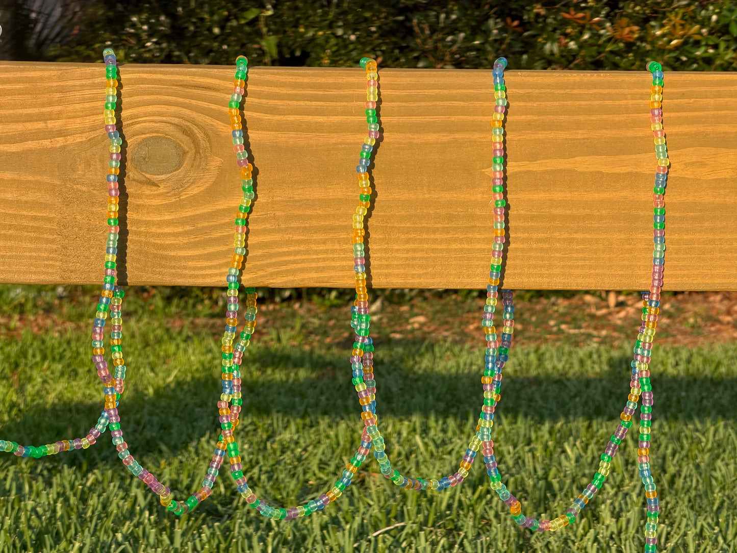 Five colorful beaded necklaces draped over a wooden fence on a sunny day, with green grass and trees in the background.