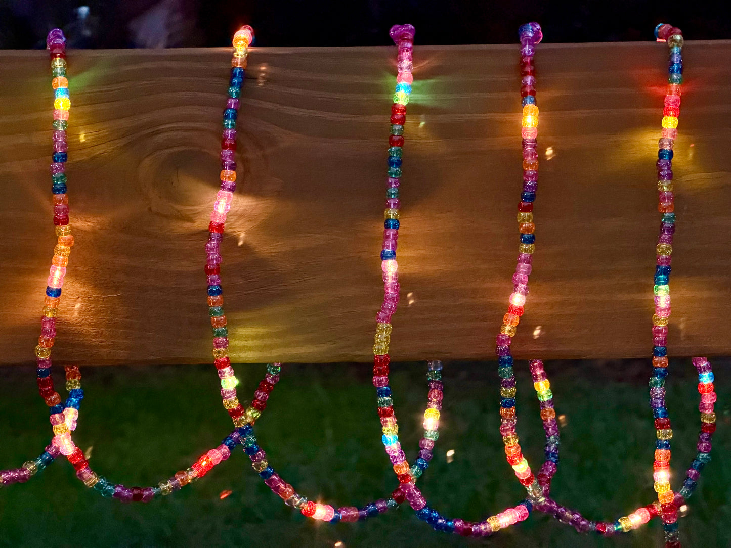 Multicolor LED beaded string lights draped over a wooden railing at night, casting warm and bright rainbow reflections on the wood and grass below.