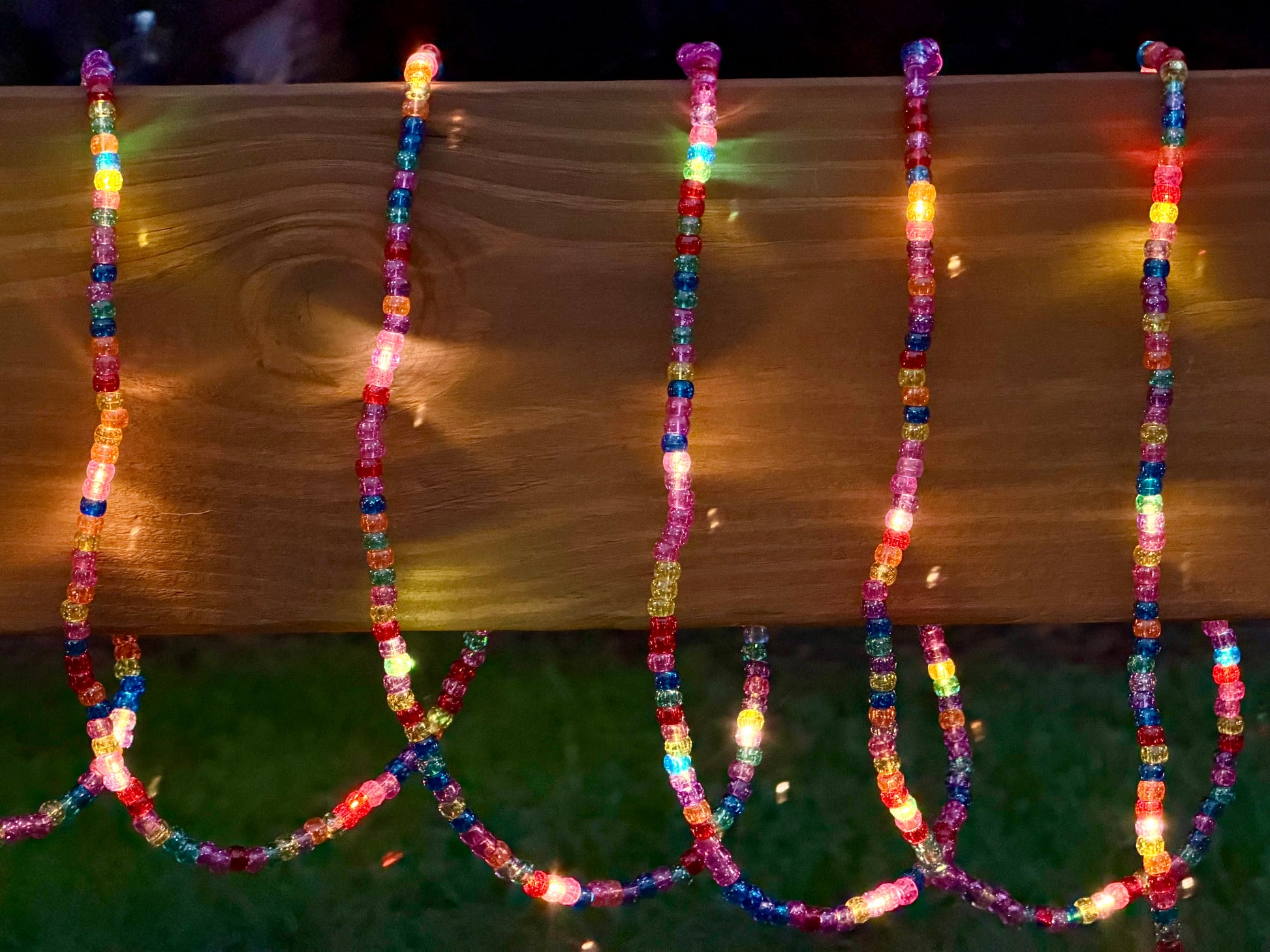 Multicolor LED beaded string lights draped over a wooden railing at night, casting warm and bright rainbow reflections on the wood and grass below.