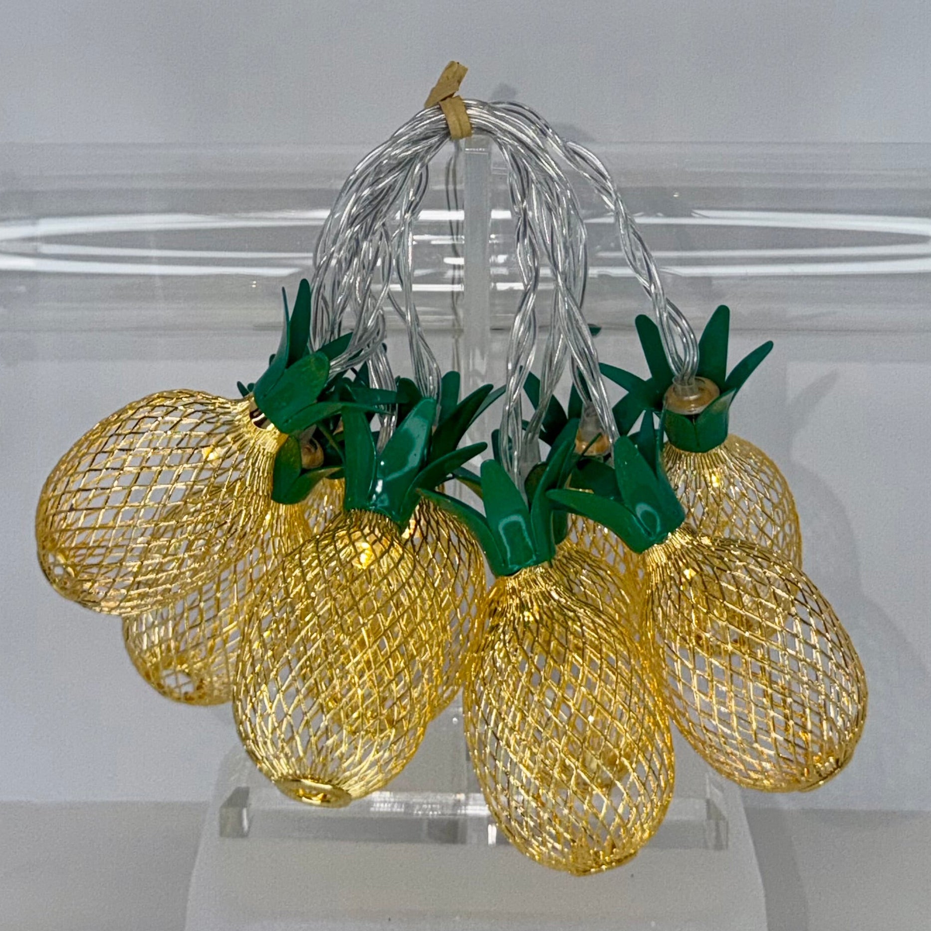 A set of pineapple-shaped string lights with glowing gold mesh bodies and green leafy tops, hanging from a clear acrylic stand in front of a transparent cylindrical display tube.