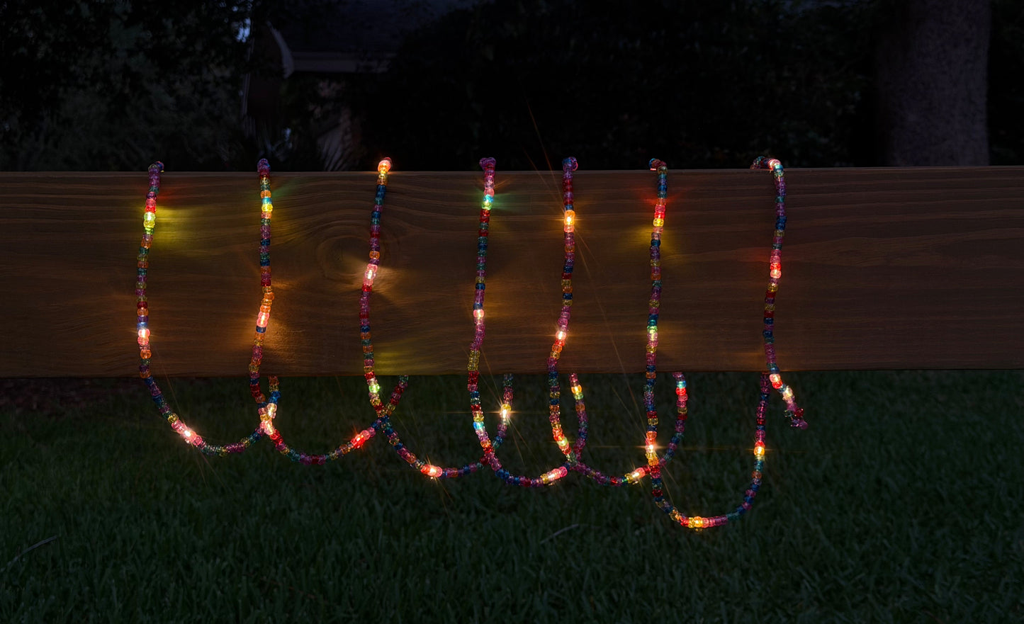 Multicolor beaded string lights draped over a wooden fence on a dark grassy lawn at dusk.