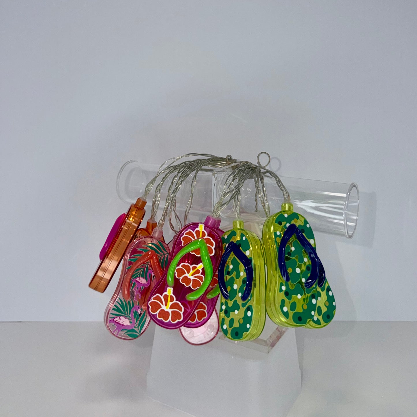 A set of six colorful flip-flop-shaped ornaments hanging from a clear plastic rod on a white display stand. The ornaments feature various tropical floral and leaf patterns in pink, orange, green, yellow, and blue.