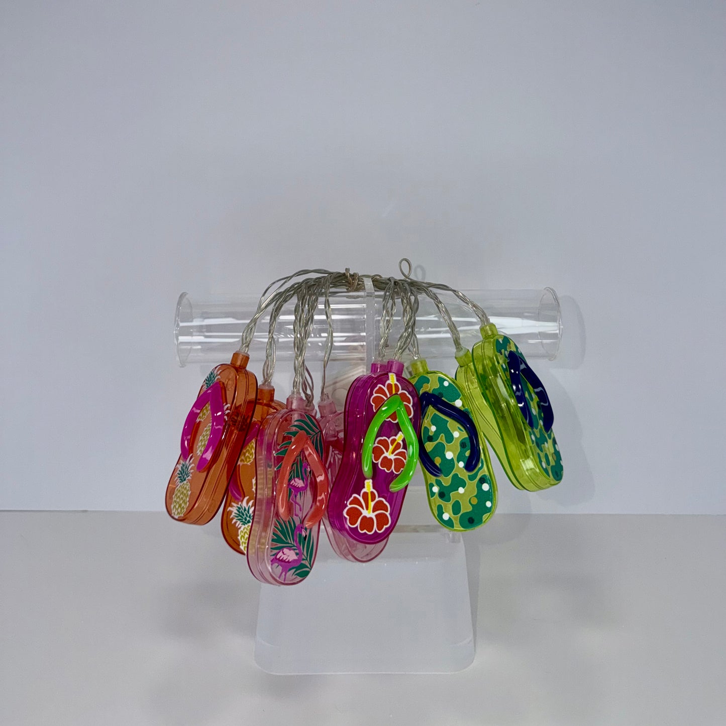A set of colorful, hand-painted glass flip-flop charms hanging from a clear plastic tube stand on a white surface against a white background.