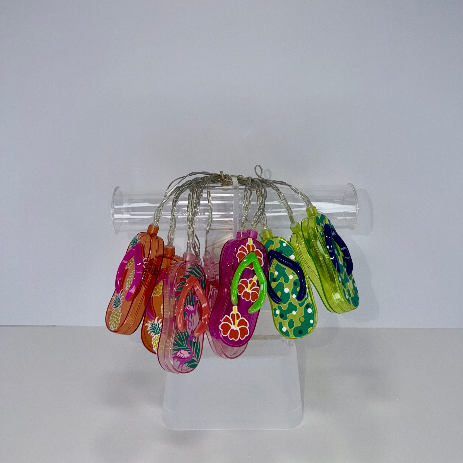 A set of colorful, hand-painted glass flip-flop charms hanging from a clear plastic tube stand on a white surface against a white background.