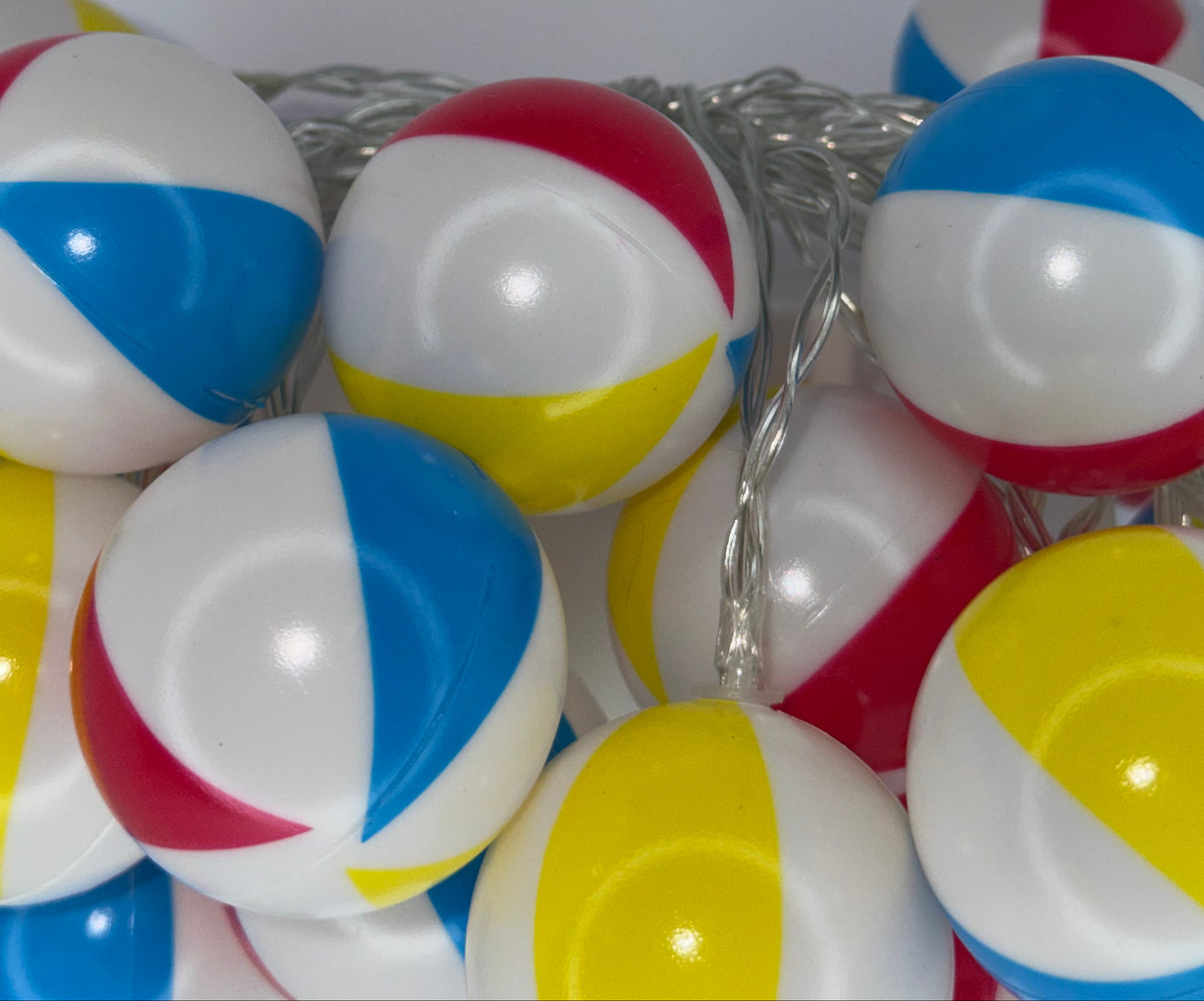 A close-up of beach ball string lights with white, blue, red, and yellow spherical bulbs wrapped in a silver wire, set against a plain light gray background.