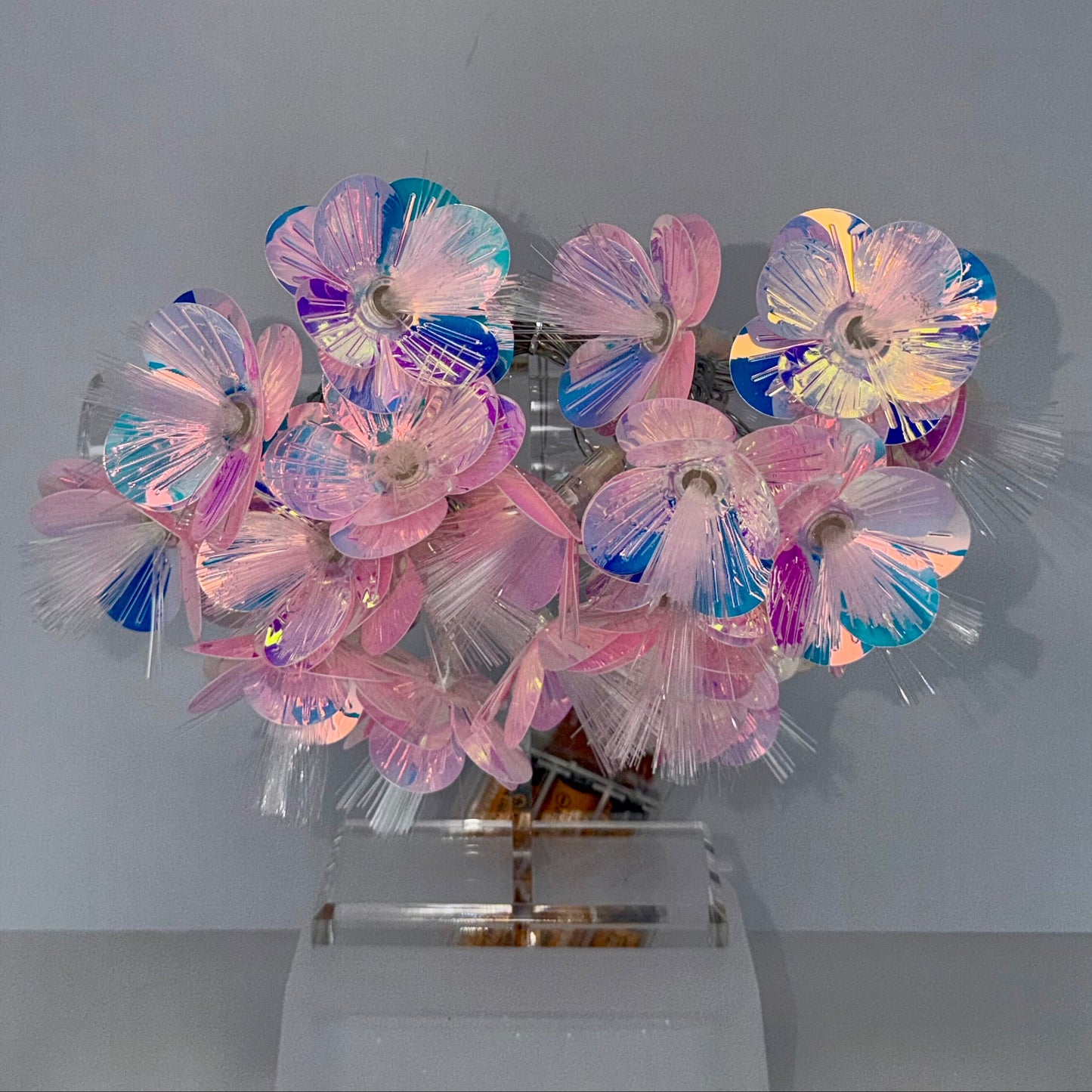 A decorative floral arrangement made of iridescent plastic flower petals in pink, blue, purple, and white, mounted on a clear acrylic stand against a plain light gray wall.