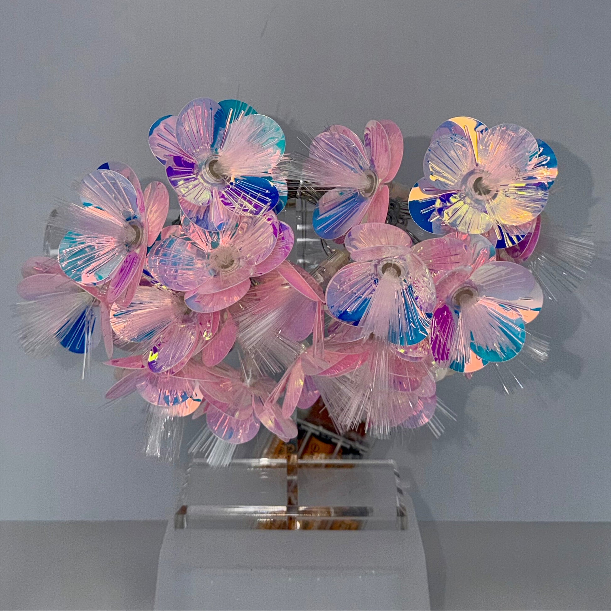 A decorative floral arrangement made of iridescent plastic flower petals in pink, blue, purple, and white, mounted on a clear acrylic stand against a plain light gray wall.