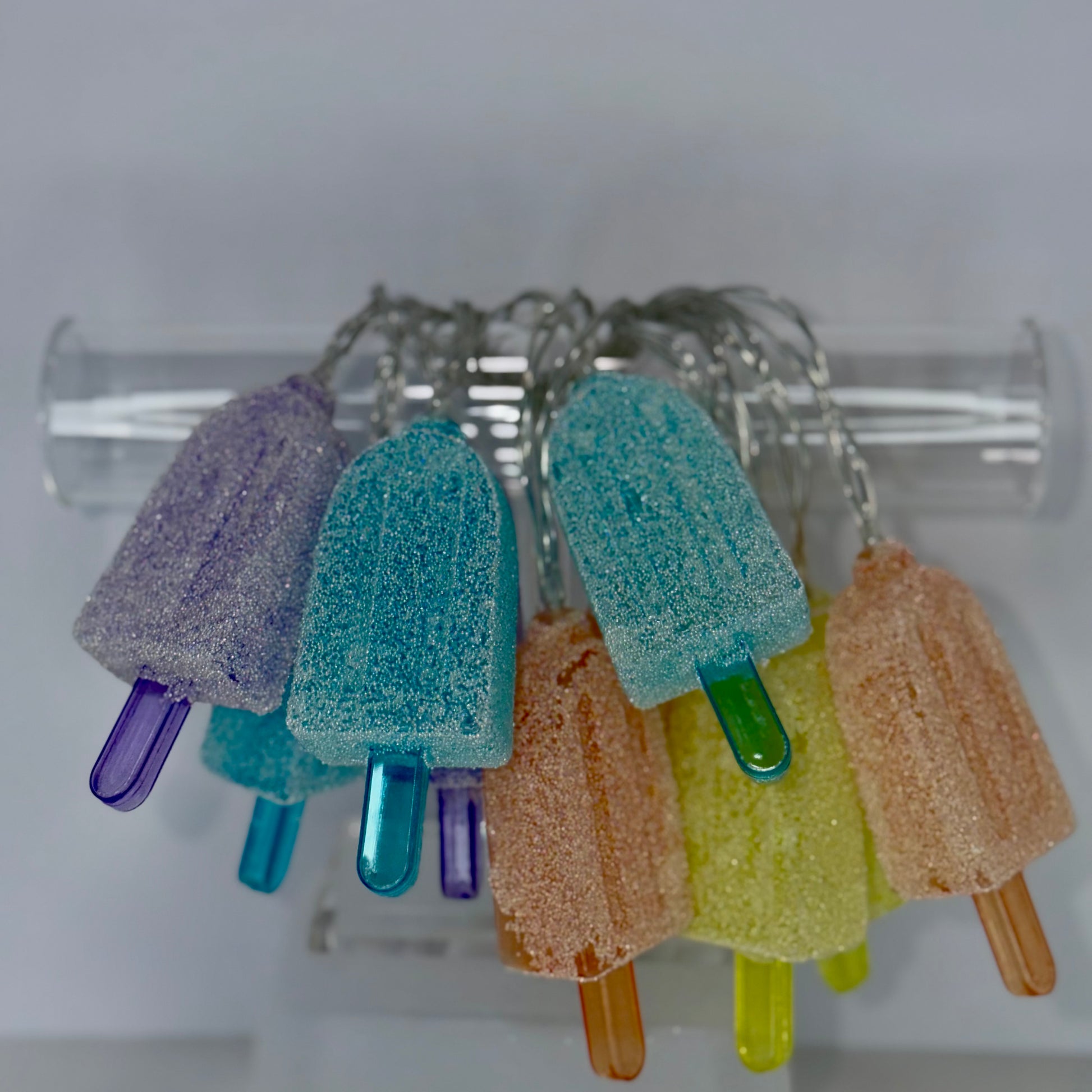A set of colorful popsicle-shaped keychains hanging from a clear plastic tube holder. The keychains come in pastel shades of purple, teal, peach, and yellow, with glittery sugar-coated surfaces and translucent plastic popsicle sticks.