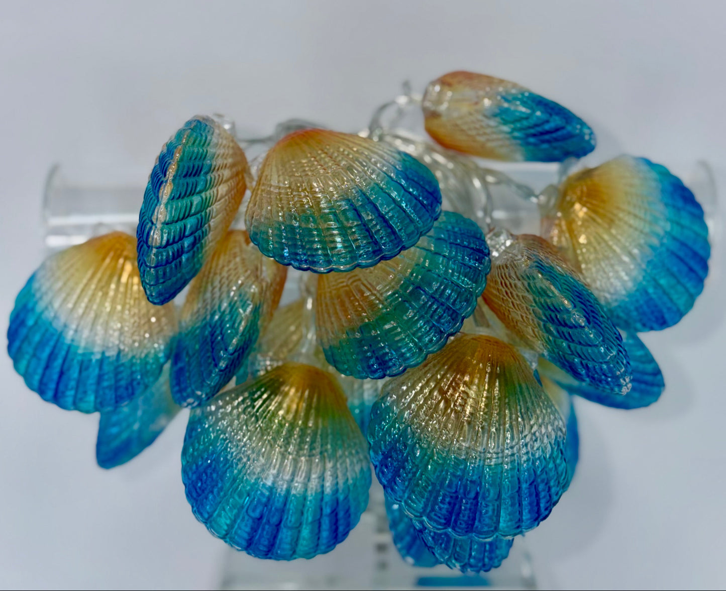 A cluster of decorative seashell-shaped ornaments or earrings with a blue-to-yellow gradient, displayed on a clear stand against a plain light background.