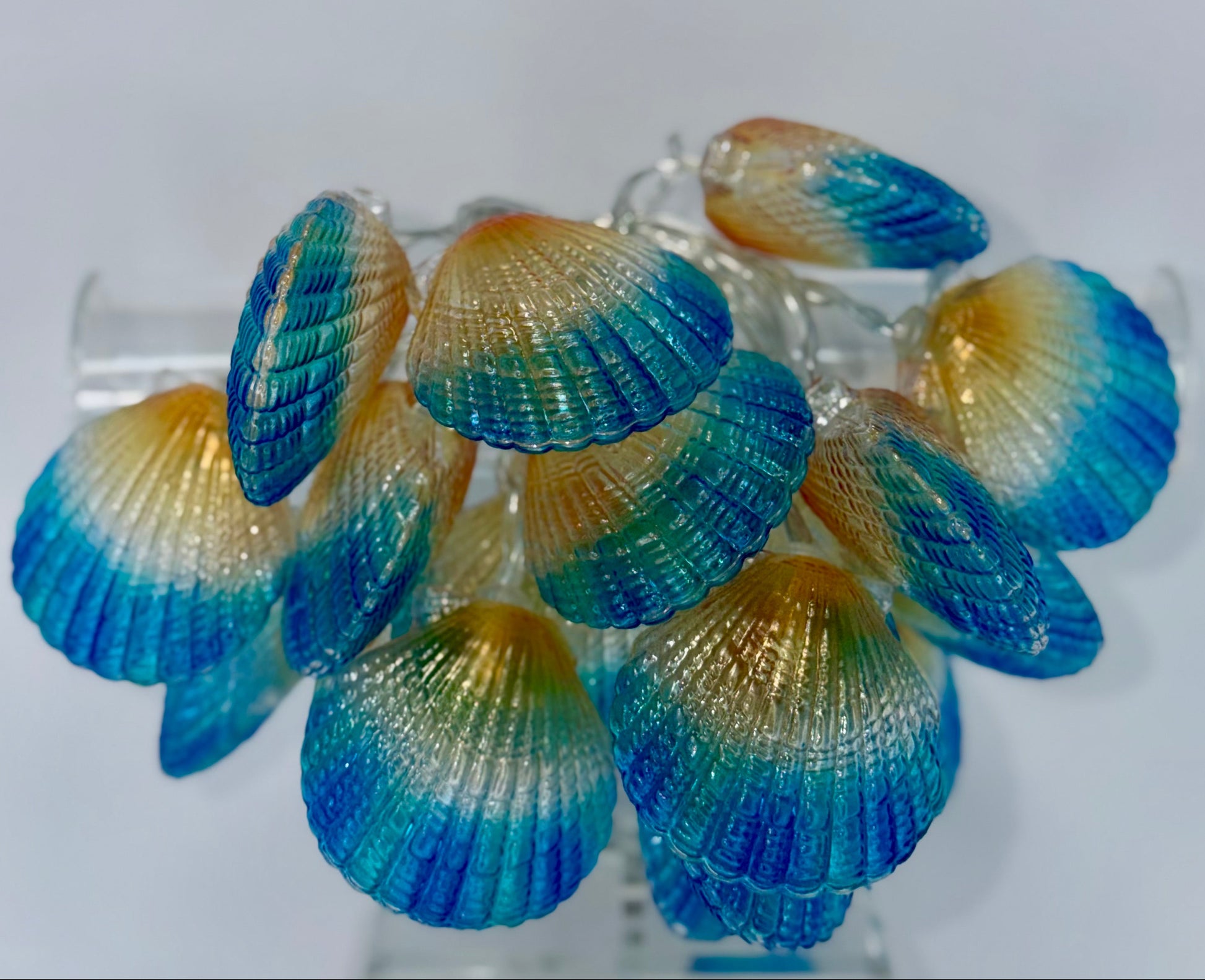 A cluster of decorative seashell-shaped ornaments or earrings with a blue-to-yellow gradient, displayed on a clear stand against a plain light background.