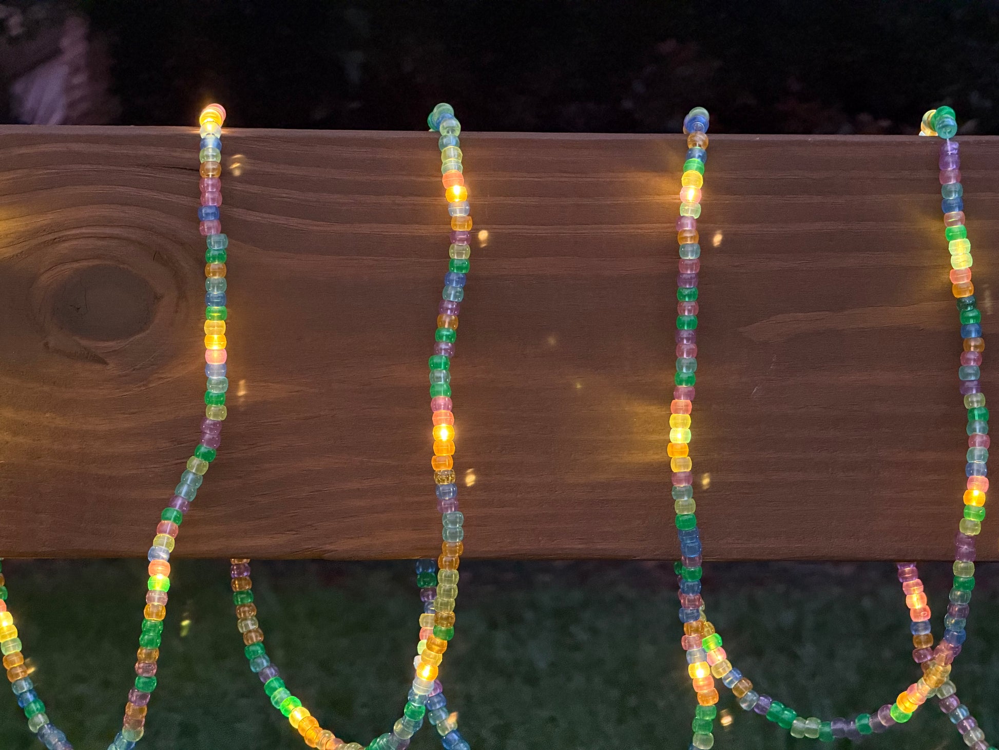 Multicolor beaded string lights draped over a wooden fence rail at night, glowing with warm yellow light.