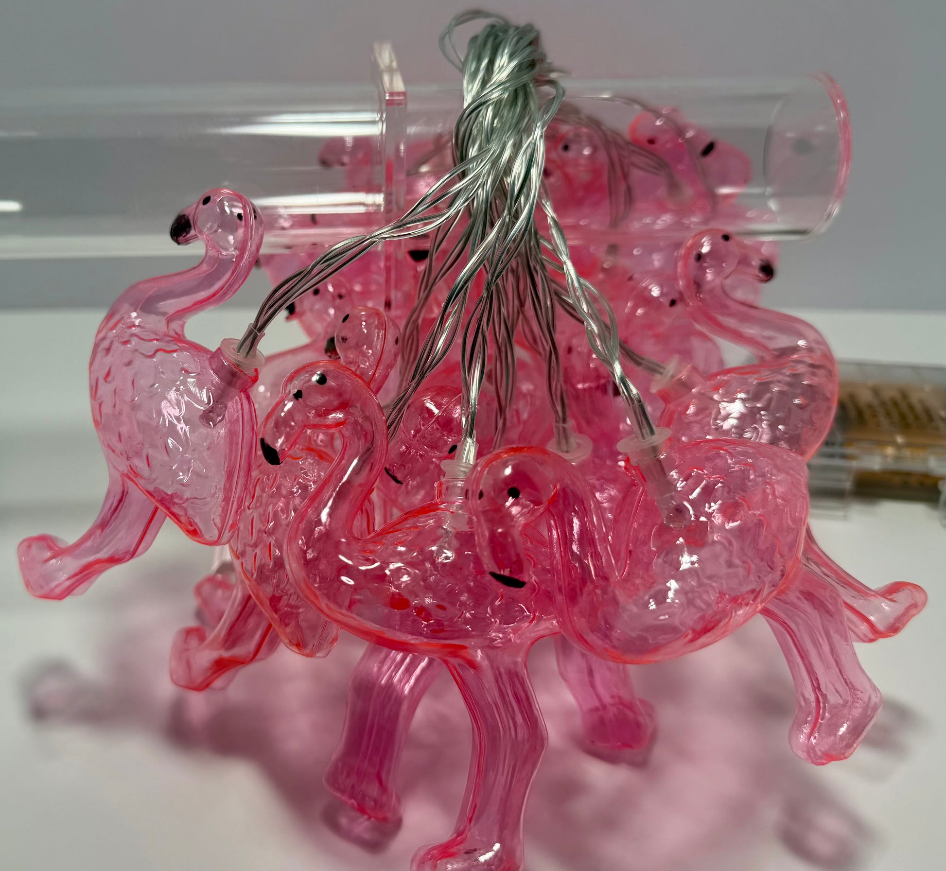 Pink translucent flamingo-shaped string lights with a silver wire, photographed on a white surface with a clear plastic tube in the background.