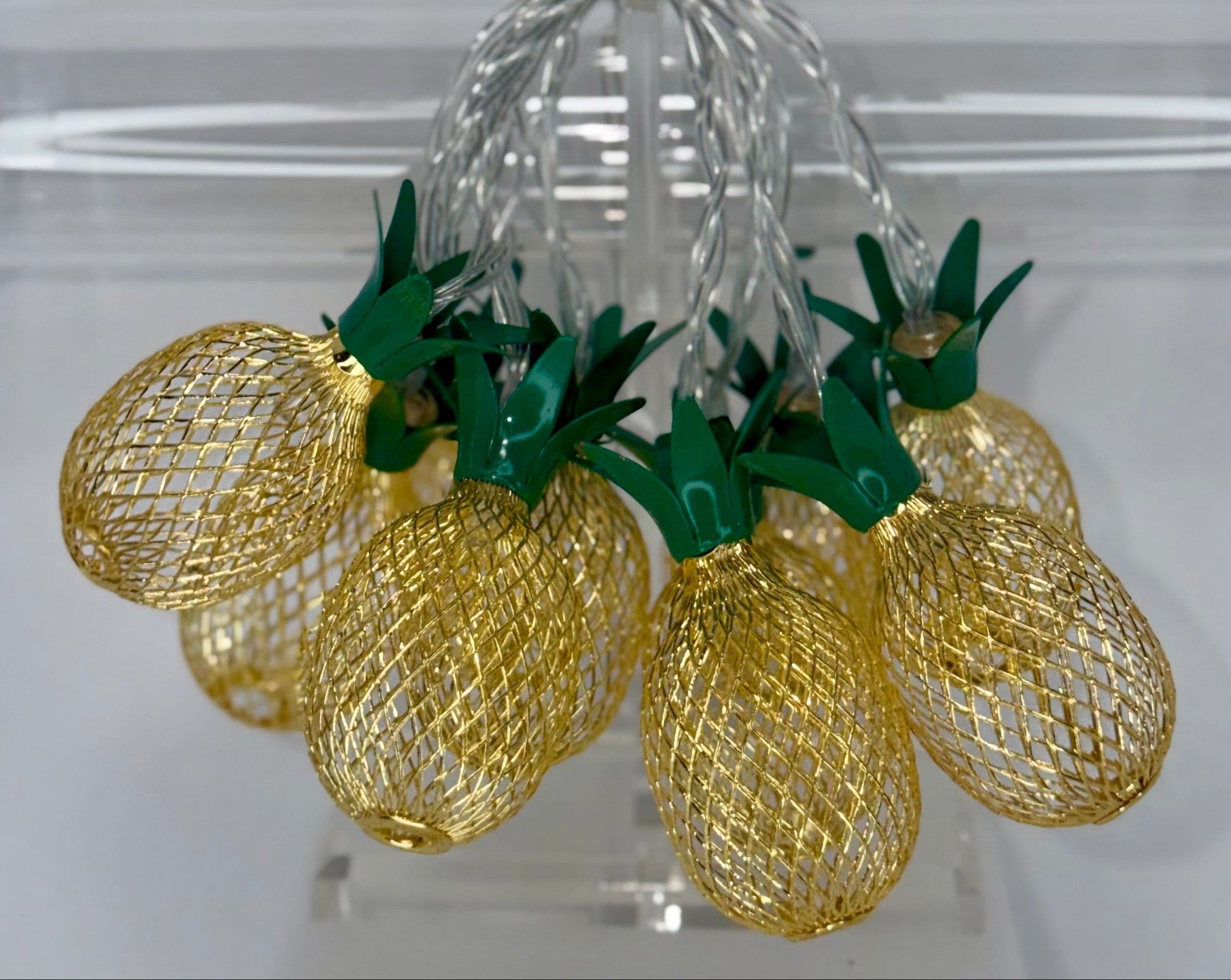 A set of decorative pineapple-shaped string lights with gold mesh bodies and green leafy tops, hanging from a clear acrylic display stand.