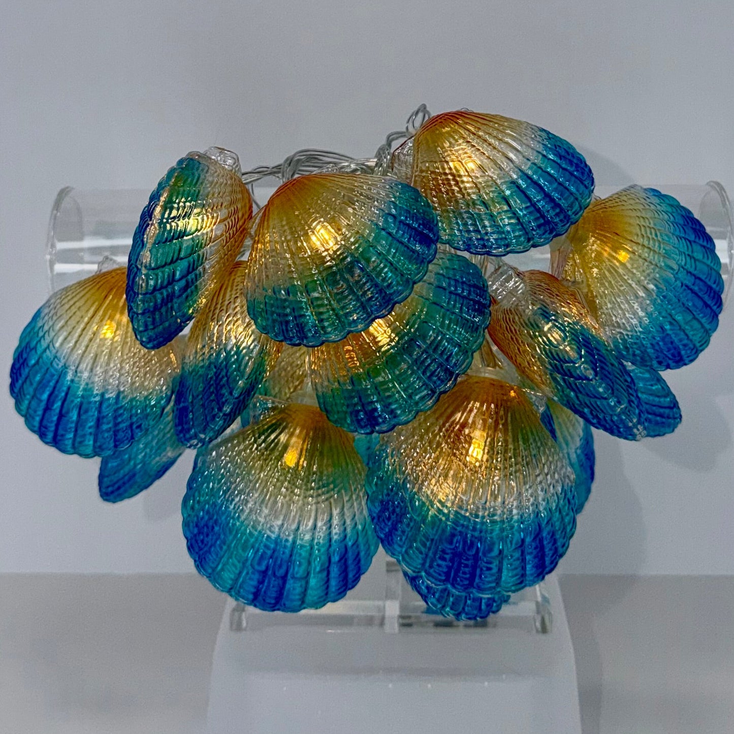 A cluster of decorative clam shell-shaped string lights with a blue-to-gold iridescent gradient, displayed on a clear acrylic stand against a plain light gray background.