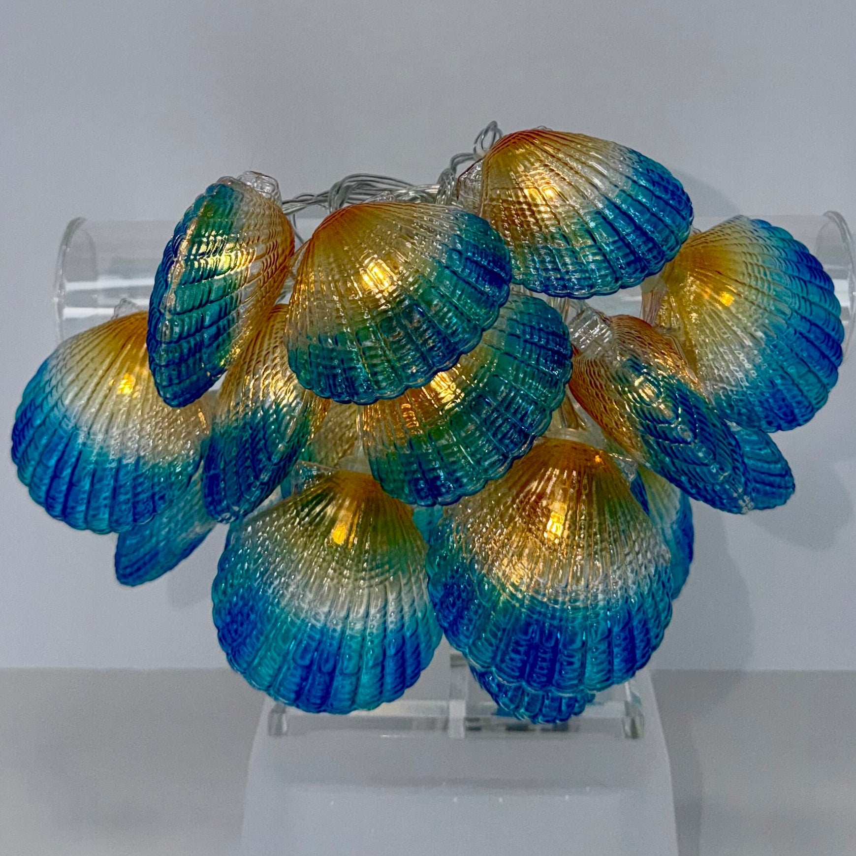 A cluster of decorative clam shell-shaped string lights with a blue-to-gold iridescent gradient, displayed on a clear acrylic stand against a plain light gray background.