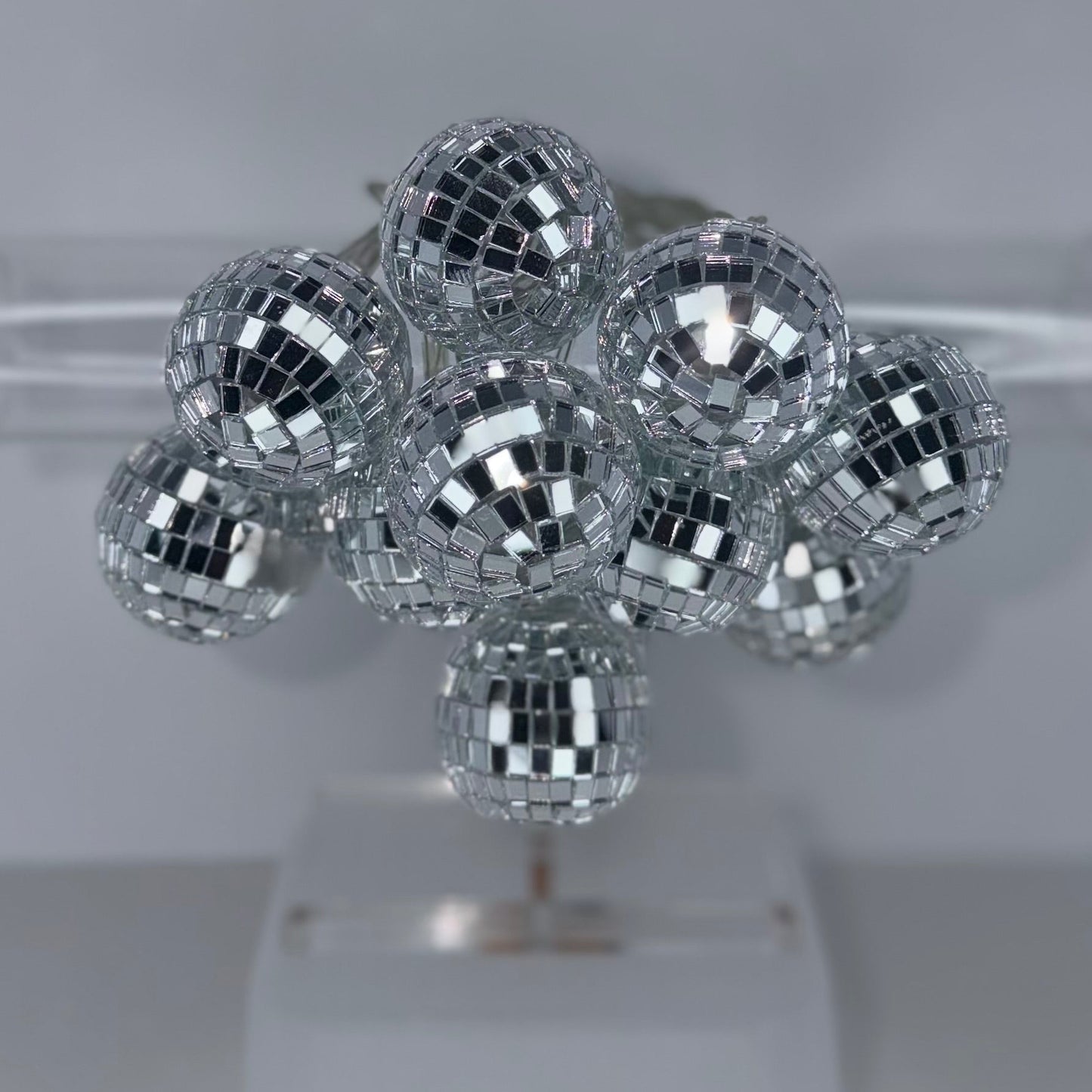 A cluster of small silver disco ball ornaments displayed on a clear stand against a light gray background.