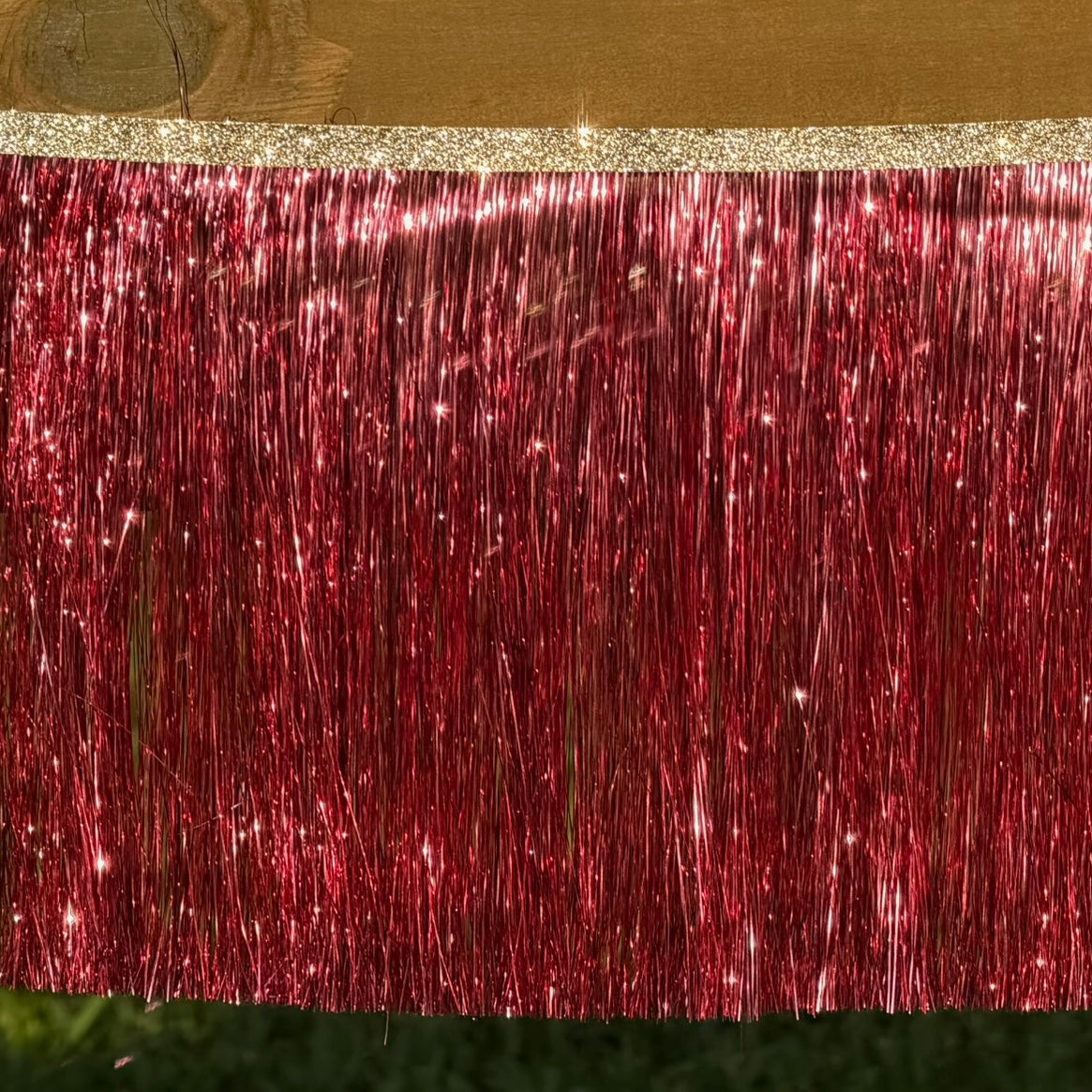 A close-up of a red metallic tinsel fringe curtain with a glittery gold top band, shown hanging against a wooden surface.
