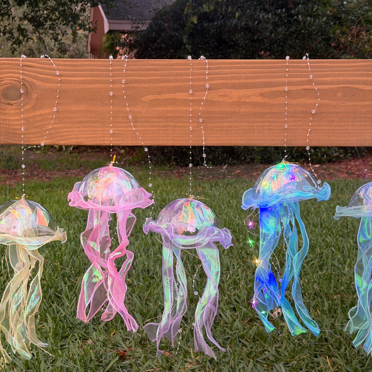 Glowing jellyfish garden lights hanging from a wooden fence on a grassy lawn at dusk, with iridescent translucent bodies and small string lights.