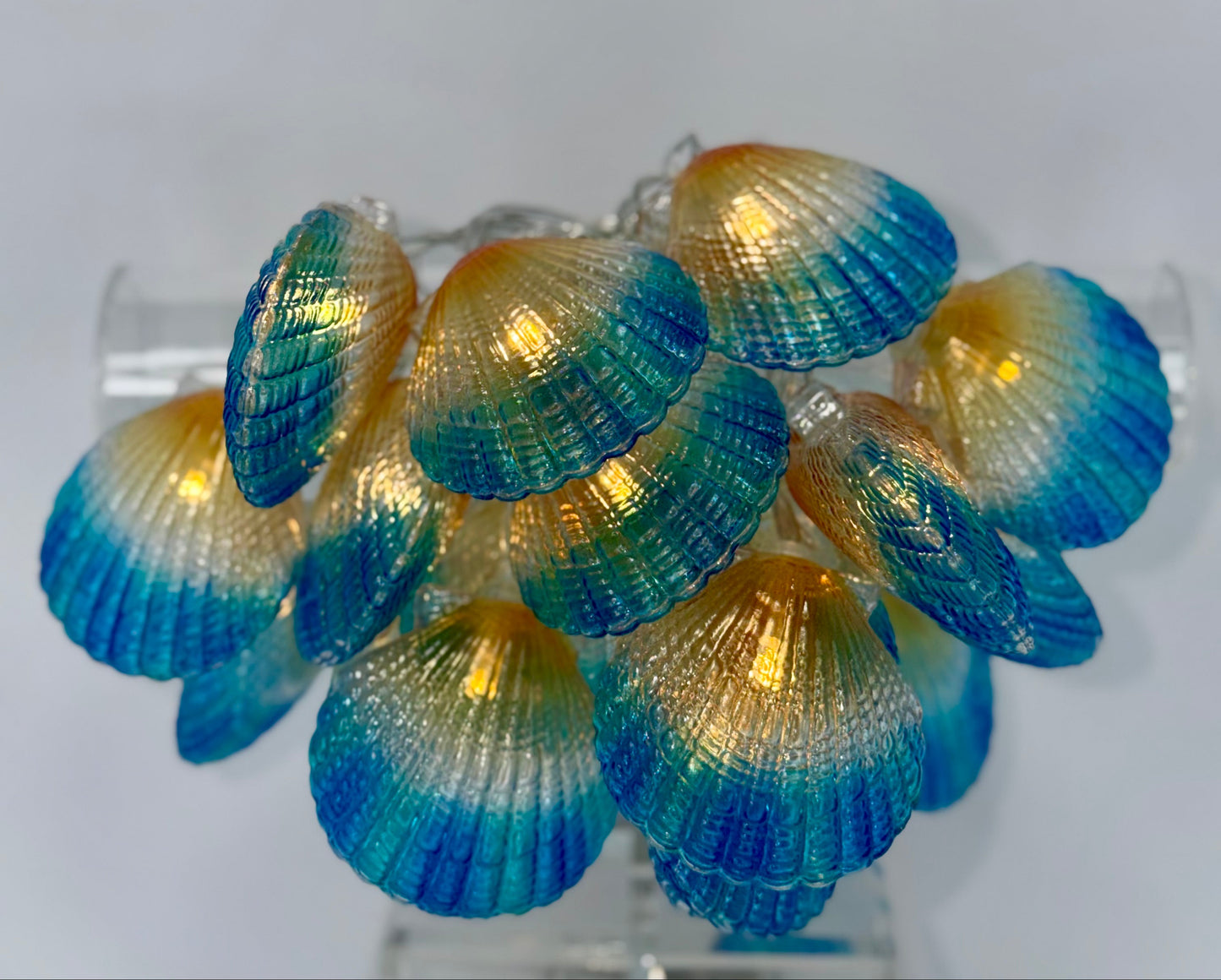 A cluster of decorative seashell-shaped lights with a blue-to-gold gradient finish, displayed on a clear stand against a light gray background.